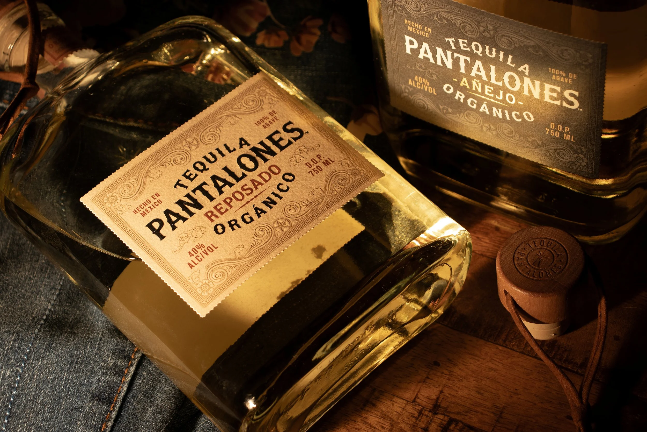 Two bottles of Tequila Pantalones Reposado, a small cork-topped bottle, and a fabric item on a wooden surface.