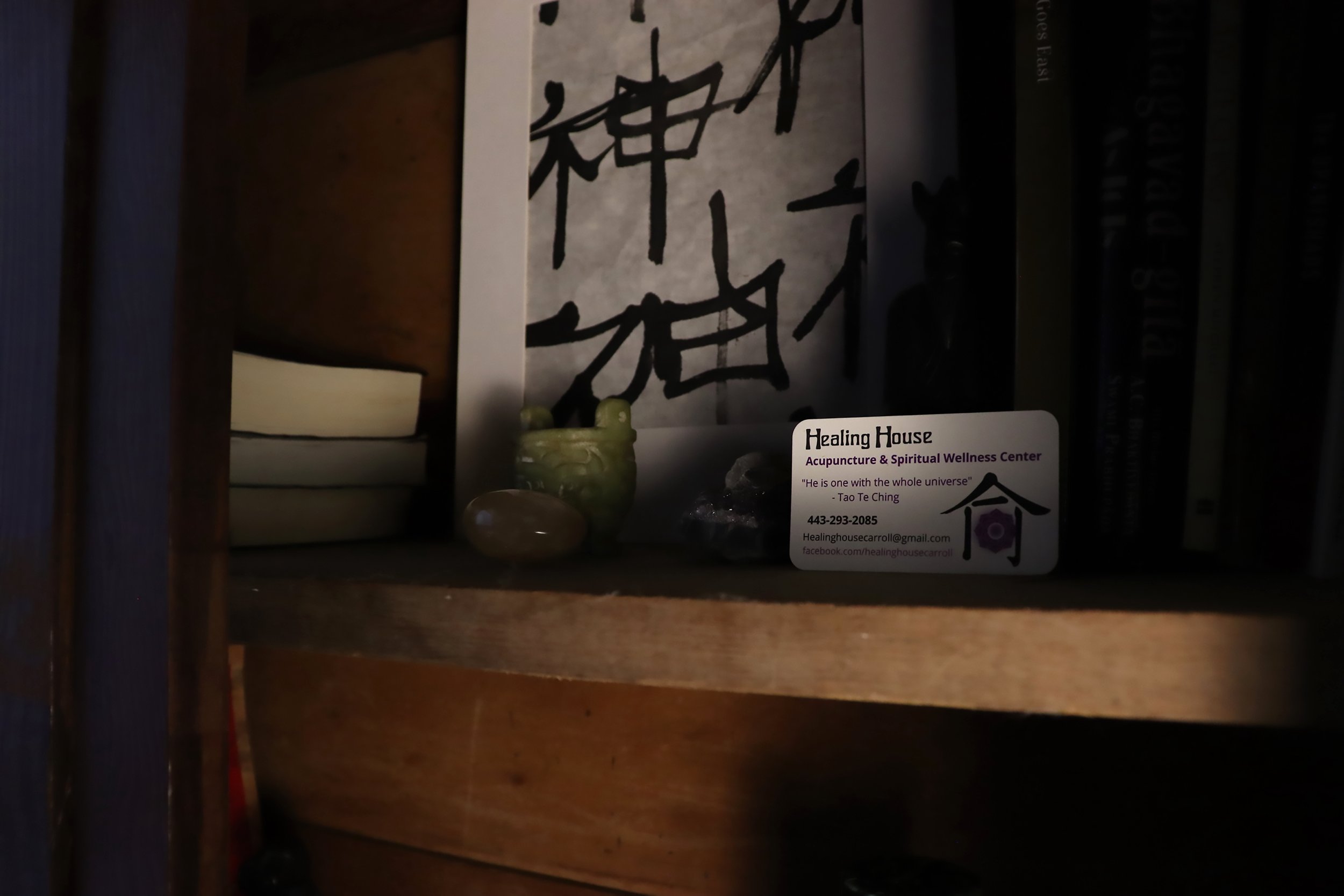 A wooden bookshelf with spiritual and wellness items, including a cactus-shaped ceramic figure, calming stones, and a business card for Healing House Acupuncture and Spiritual Wellness Center, with a background featuring a canvas with abstract black