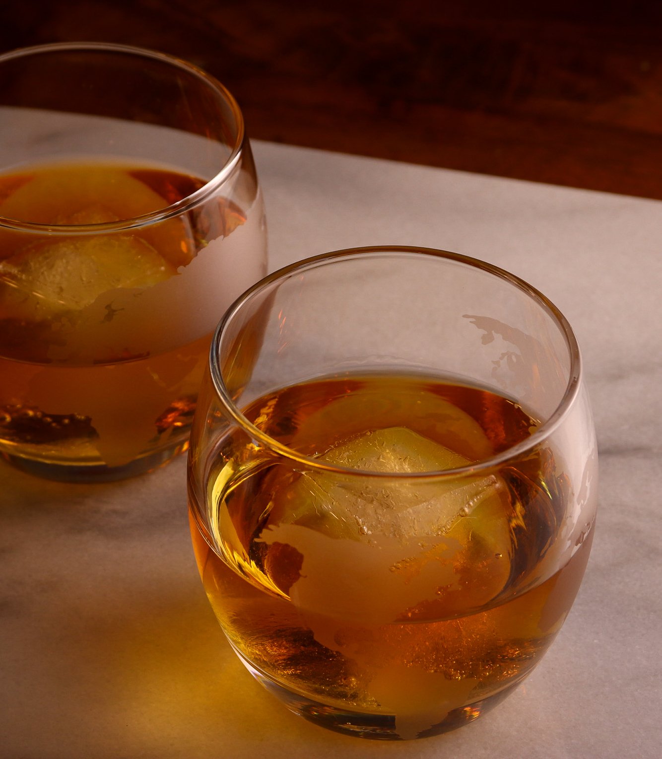 Two glasses filled with whiskey and ice cubes, on a marble surface.