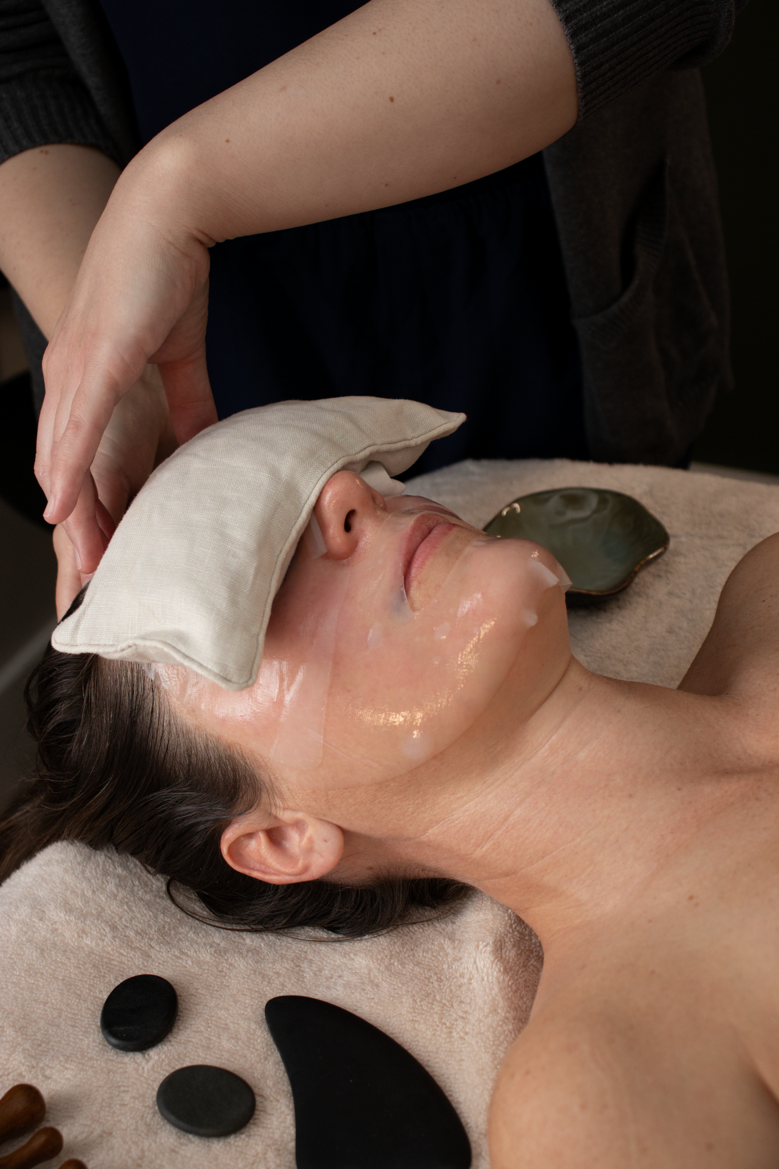 A woman receiving a facial treatment with a facial mask, lying on a spa bed with stones around her for decoration.