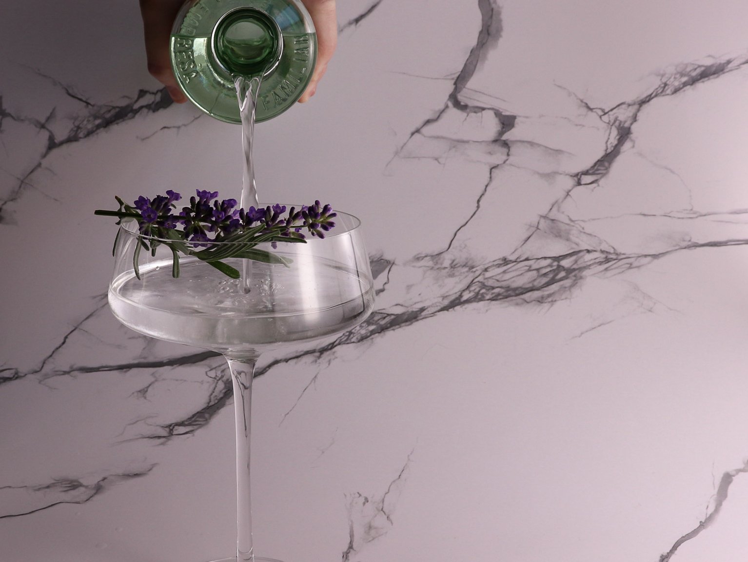 A hand pouring water from a green bottle into a wide glass with purple flowers inside, against a white marble background.