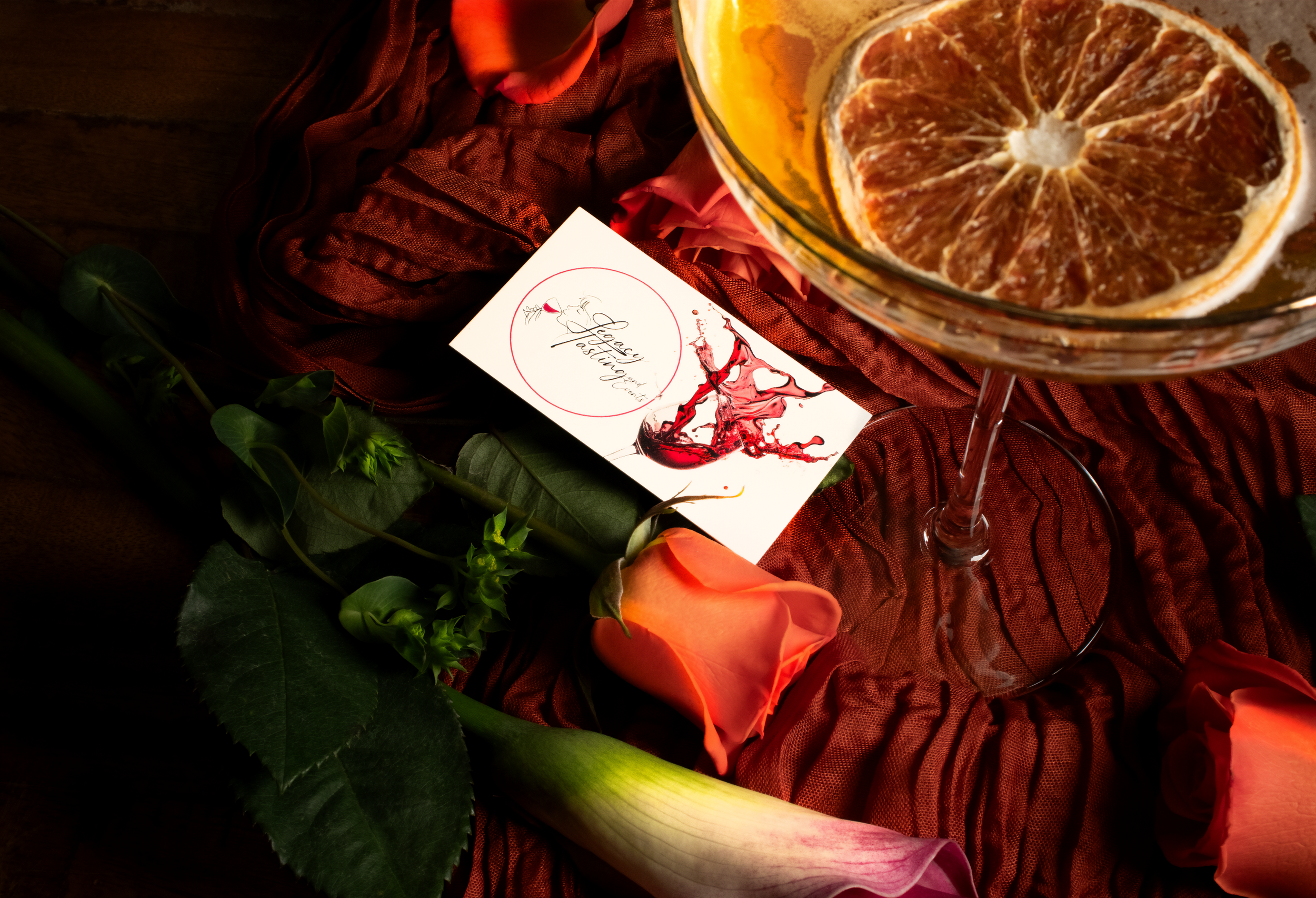 A glass filled with a beverage garnished with a dried orange slice, a pink rose, green leaves, and an unlit candle on a textured red cloth, with a card that reads "Legacy Tasting" in elegant handwriting. Photographed by Drink Flow Media Studio