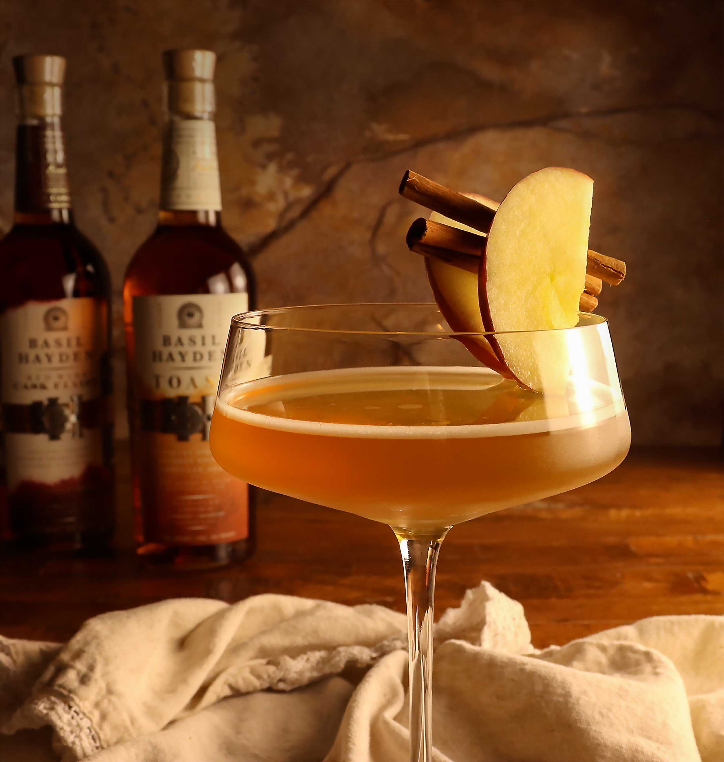 A cocktail garnished with apple slices and cinnamon sticks on a wooden surface with two bottles of Basil Hayden's bourbon in the background