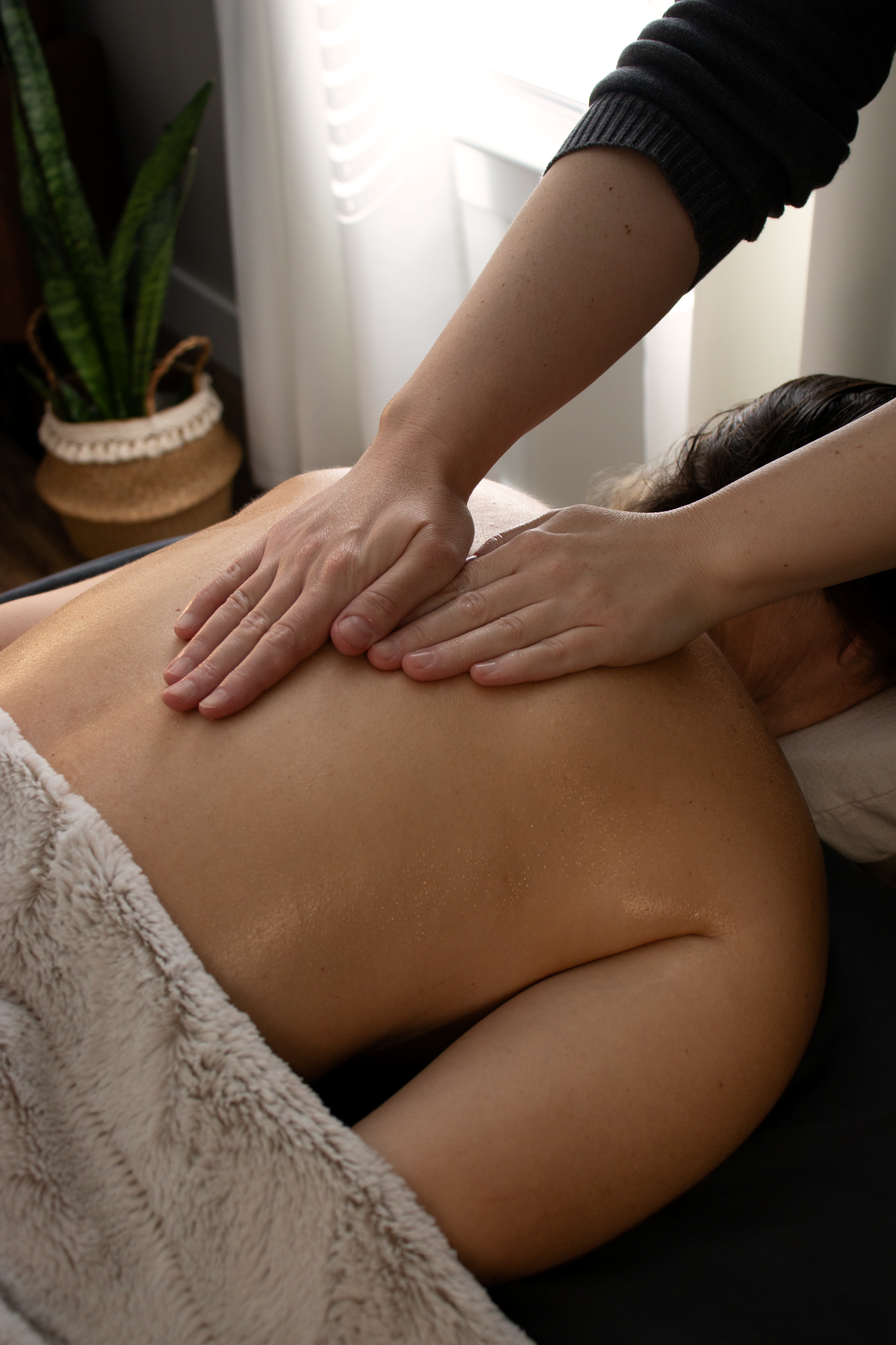 A person receiving a massage on their back in a cozy, well-lit room with a potted plant and sheer curtains.