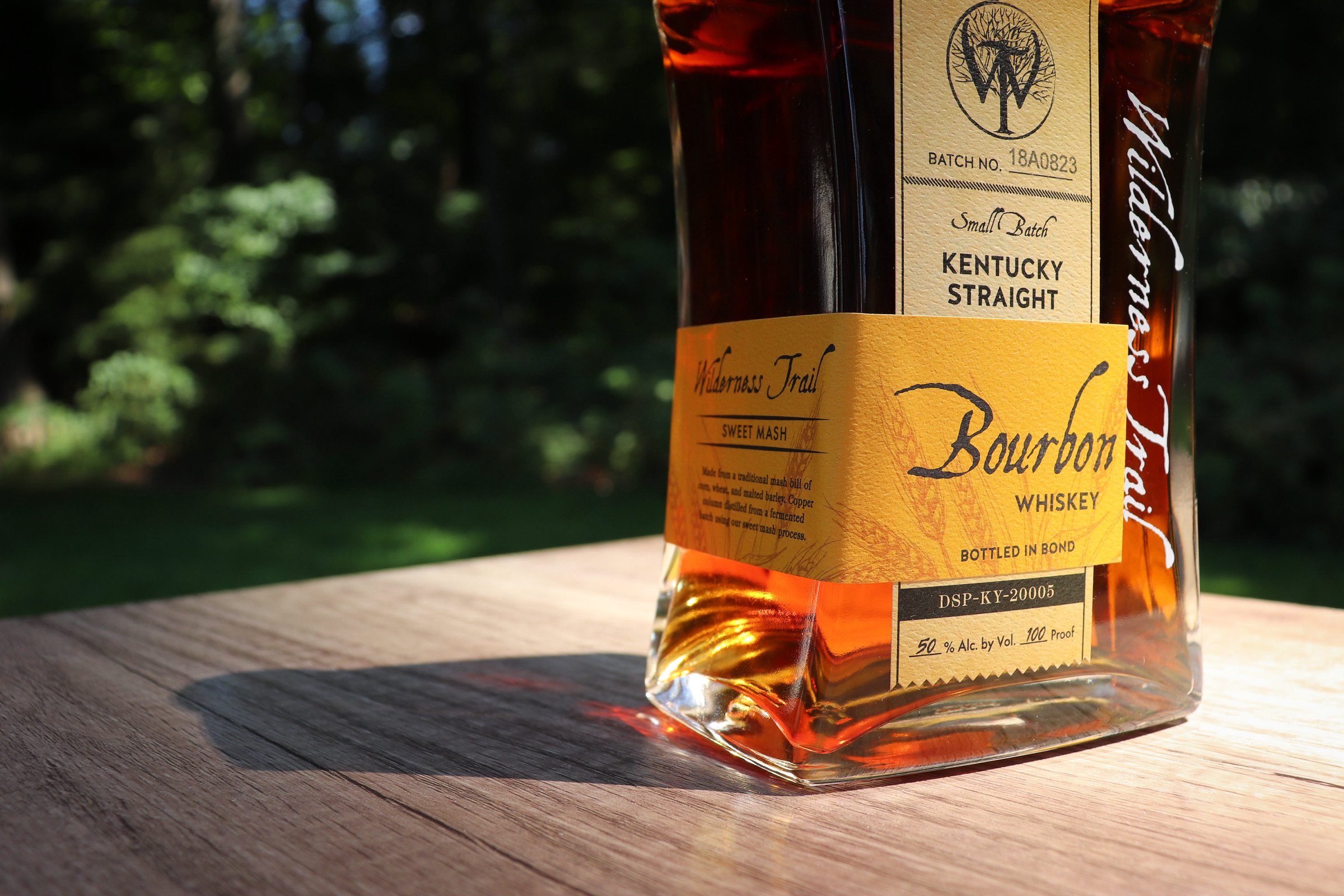 A bottle of Bourbon whiskey placed on a wooden surface outdoors, with a blurred green natural background. The label indicates it is Kentucky Straight, bottled in bond, 50% alcohol by volume.