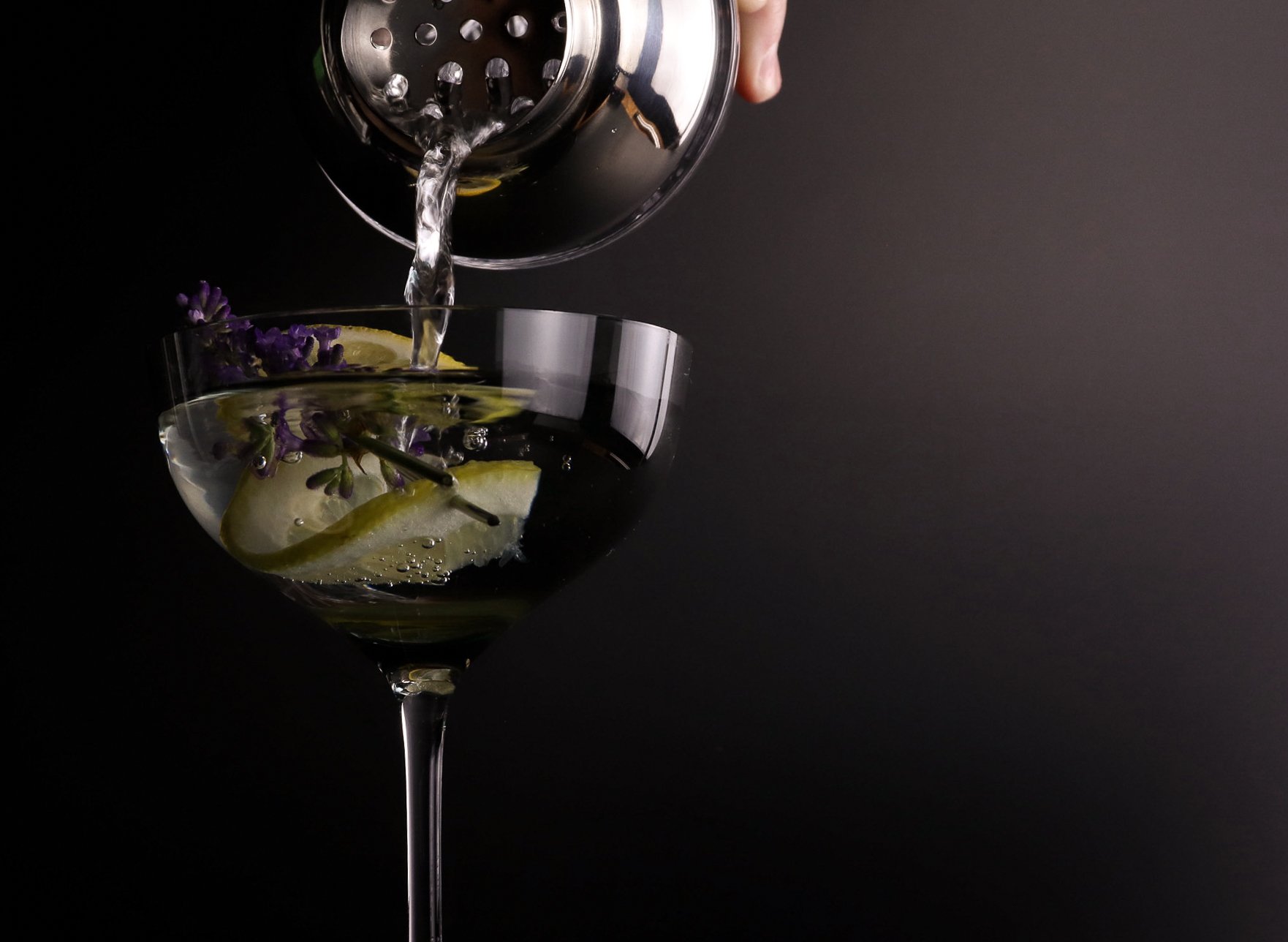 A hand pouring clear liquid into a cocktail glass with lemon slices and purple flowers, set against a dark background.