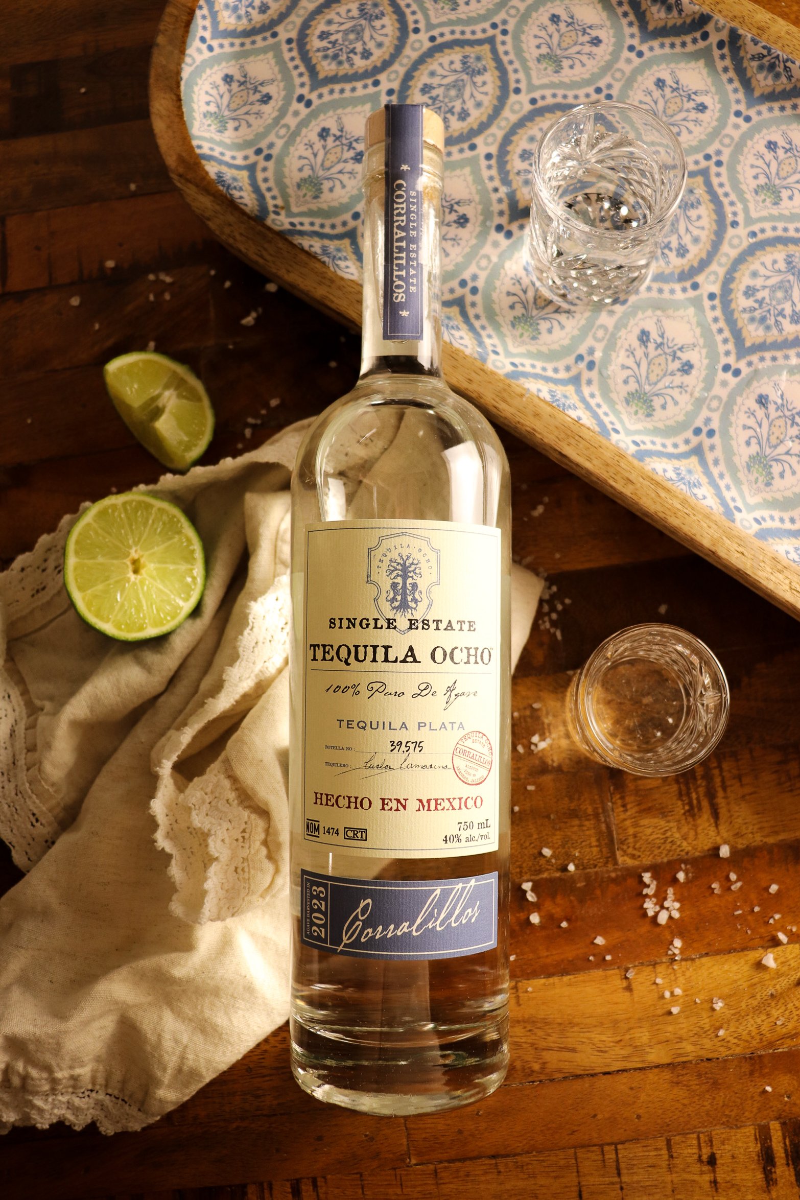 A bottle of tequila Ocho single estate on a wooden table, surrounded by two lime halves, two glasses filled with tequila, and a cloth, with salt spilled on the table edge.