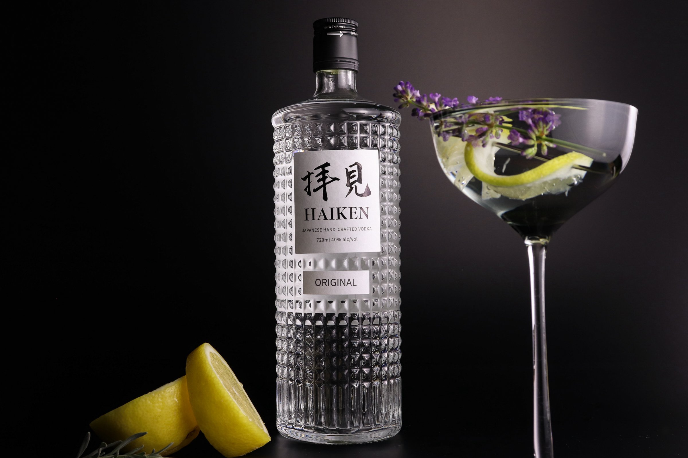 A bottle of Haiken Japanese handcrafted vodka with a textured glass surface, a martini glass with lemon wedge and purple flowers, and lemon wedges in front, all against a black background.