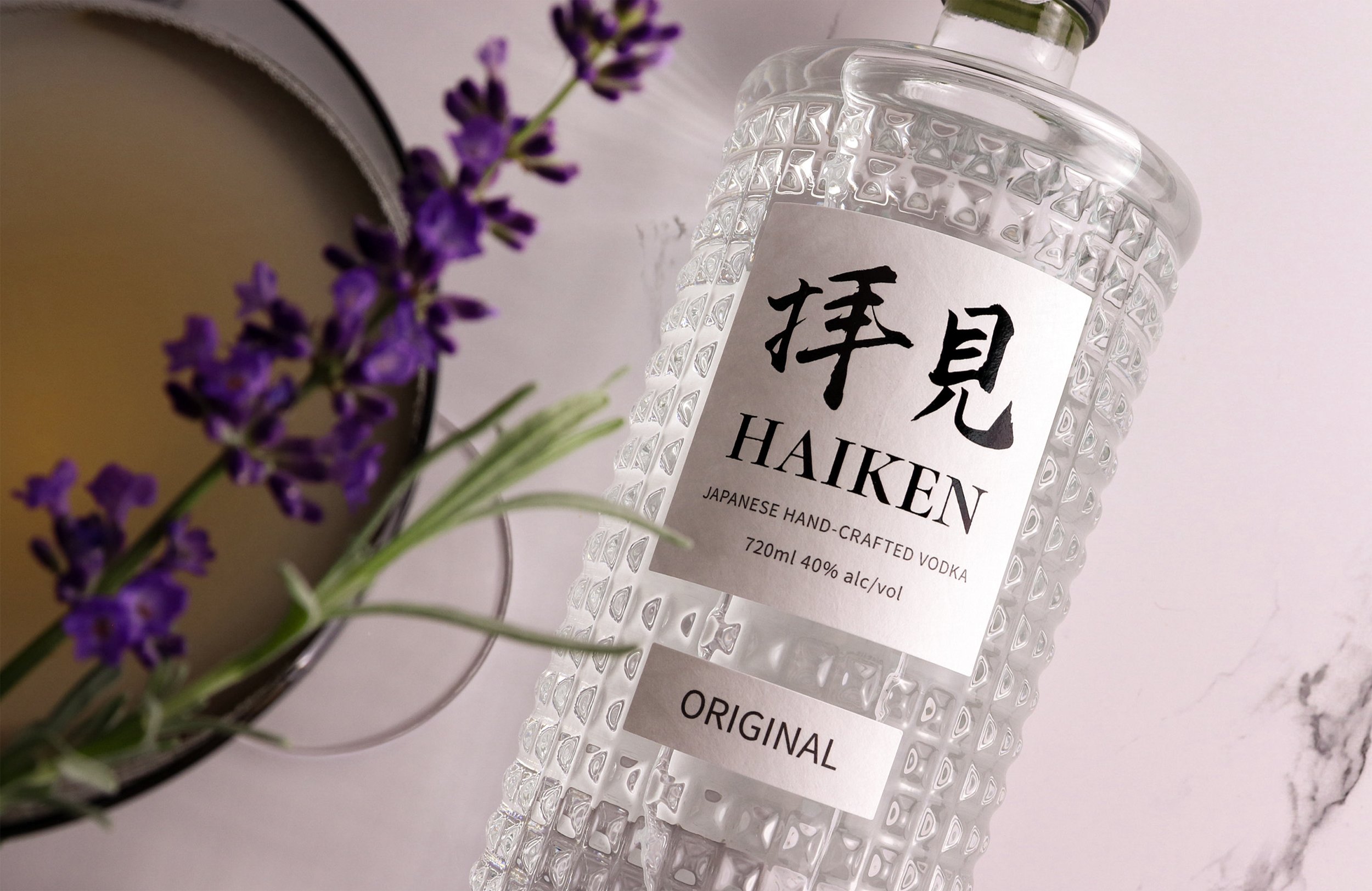 A clear textured bottle of Japanese handcrafted vodka labeled 'Haiken' with Japanese characters, placed on a light surface next to a purple lavender flower and a container with a yellowish liquid.