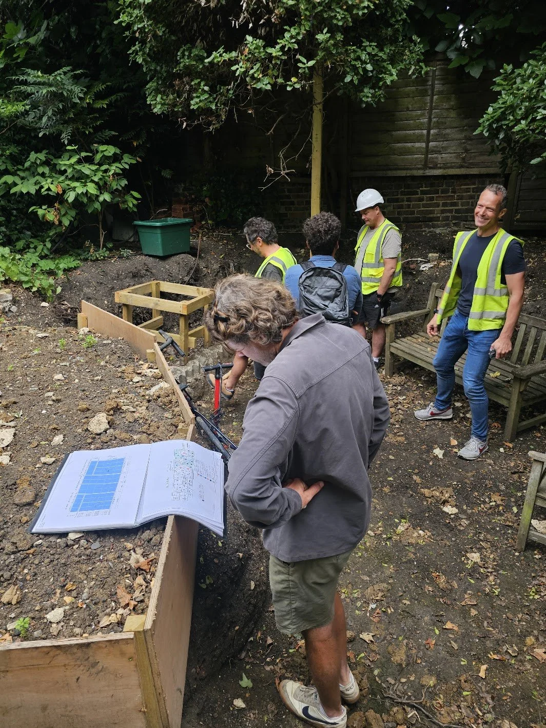 James Innes, architects, and landscape team at a project construction site, some wearing safety vests and helmets, examining foundation or groundwork in a backyard.