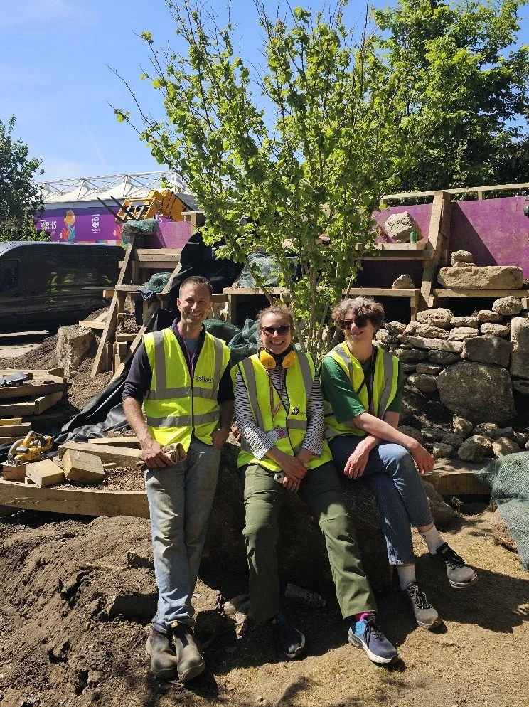 Garden designers, including James Innes, working at the RHS Chelsea Flower Show in yellow safety vests sitting and standing in front of a young tree and a rock wall during a construction or landscaping project on a sunny day.