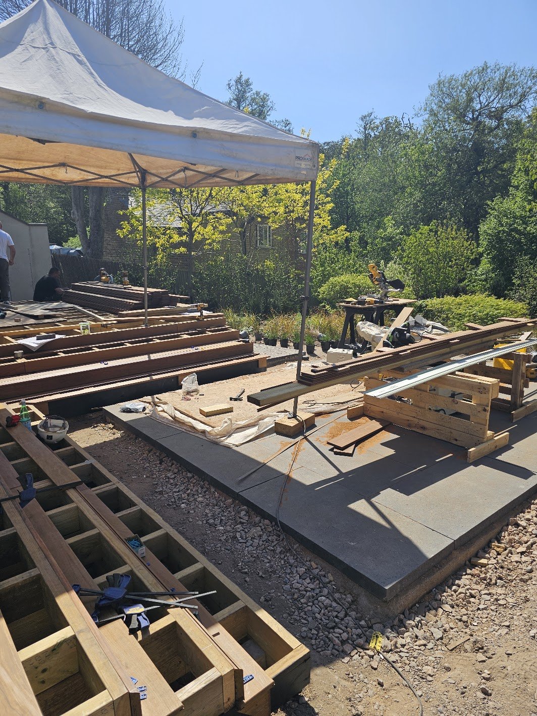Construction site outdoors  on a James Innes garden project with wooden framing and tools, working on building a bespoke hardwood deck and patio, under a white canopy, surrounded by trees and blue sky.
