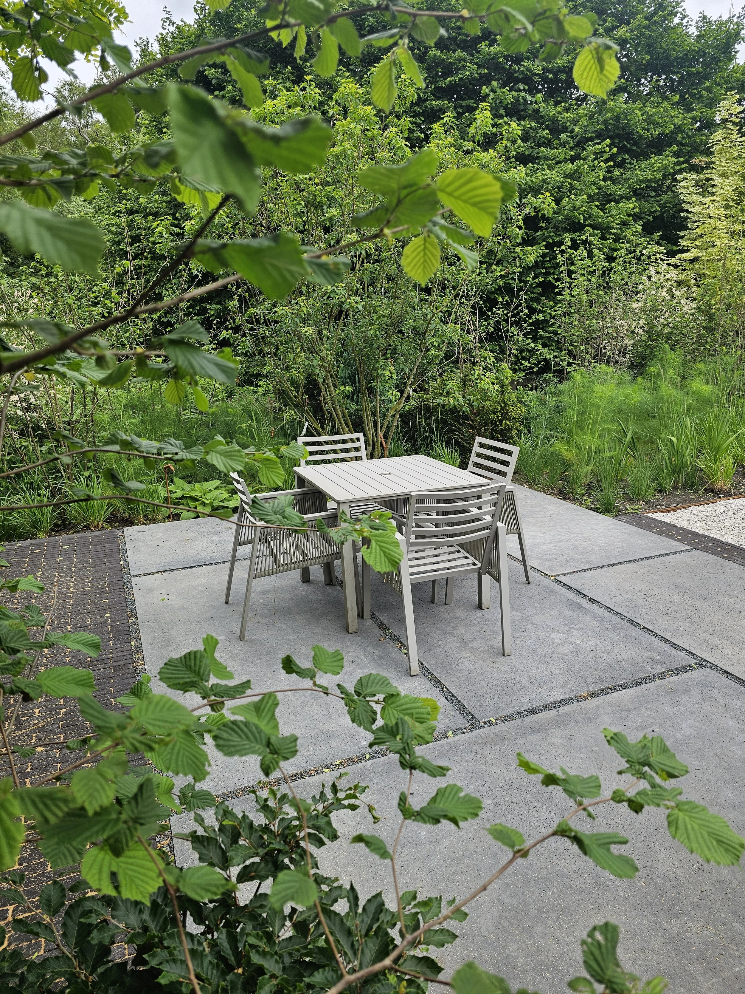 A contemporary seating area with architectural concrete slabs and dark charcoal clay pavers set within lush forest glade style planting in Virginia Water, Surrey.