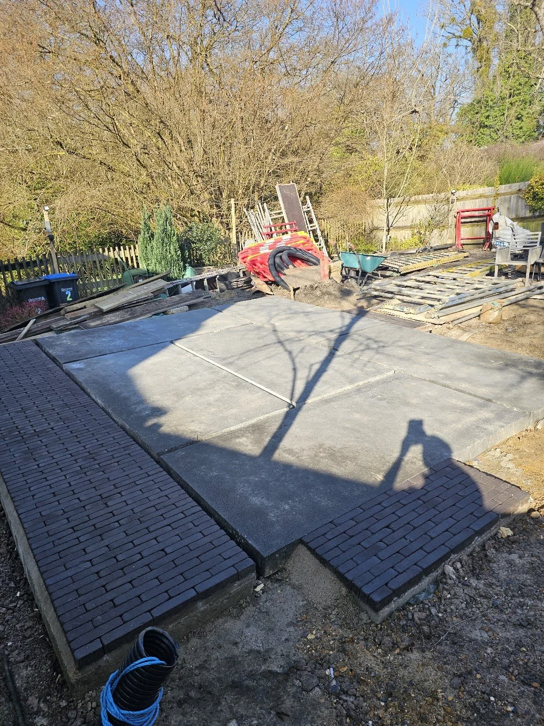 A garden in the process of being renovated with construction materials and tools scattered around, a concrete contemporary seating area, and a patio edge with clay pavers.