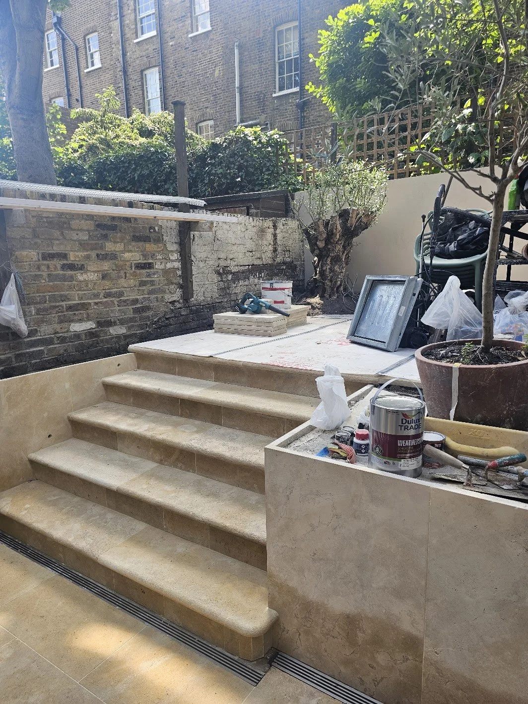 Outdoor terrace area under construction with beige limestone stone steps and flooring, construction tools, paint cans, and gardening supplies. Potted trees and bushes are visible in the background in this Islington garden.