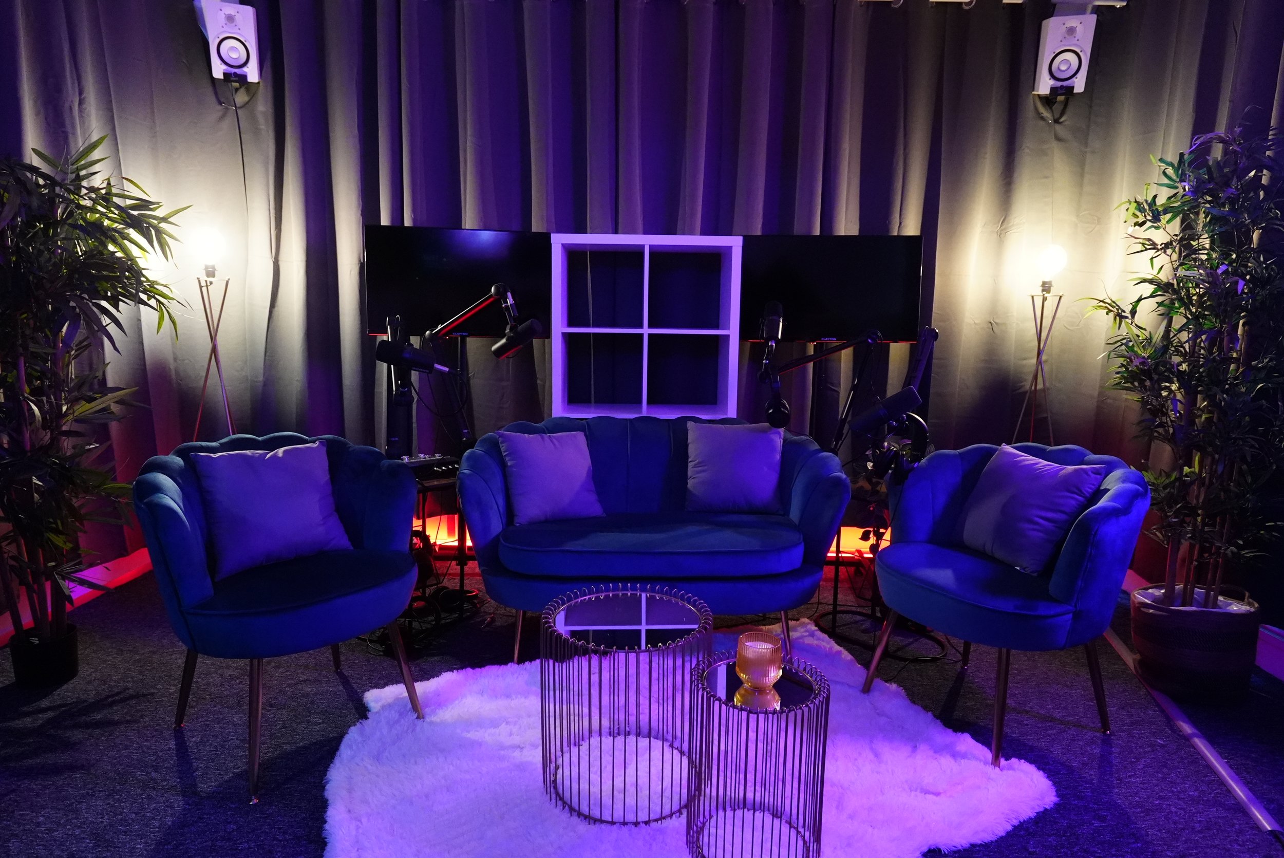 Un salon décoré avec un canapé et deux fauteuils bleus, un tapis blanc, deux tables rondes métalliques, une télévision à l'arrière, des lampes sur pied, des plantes vertes et des micros suspendus, dans un éclairage violet et chaud.