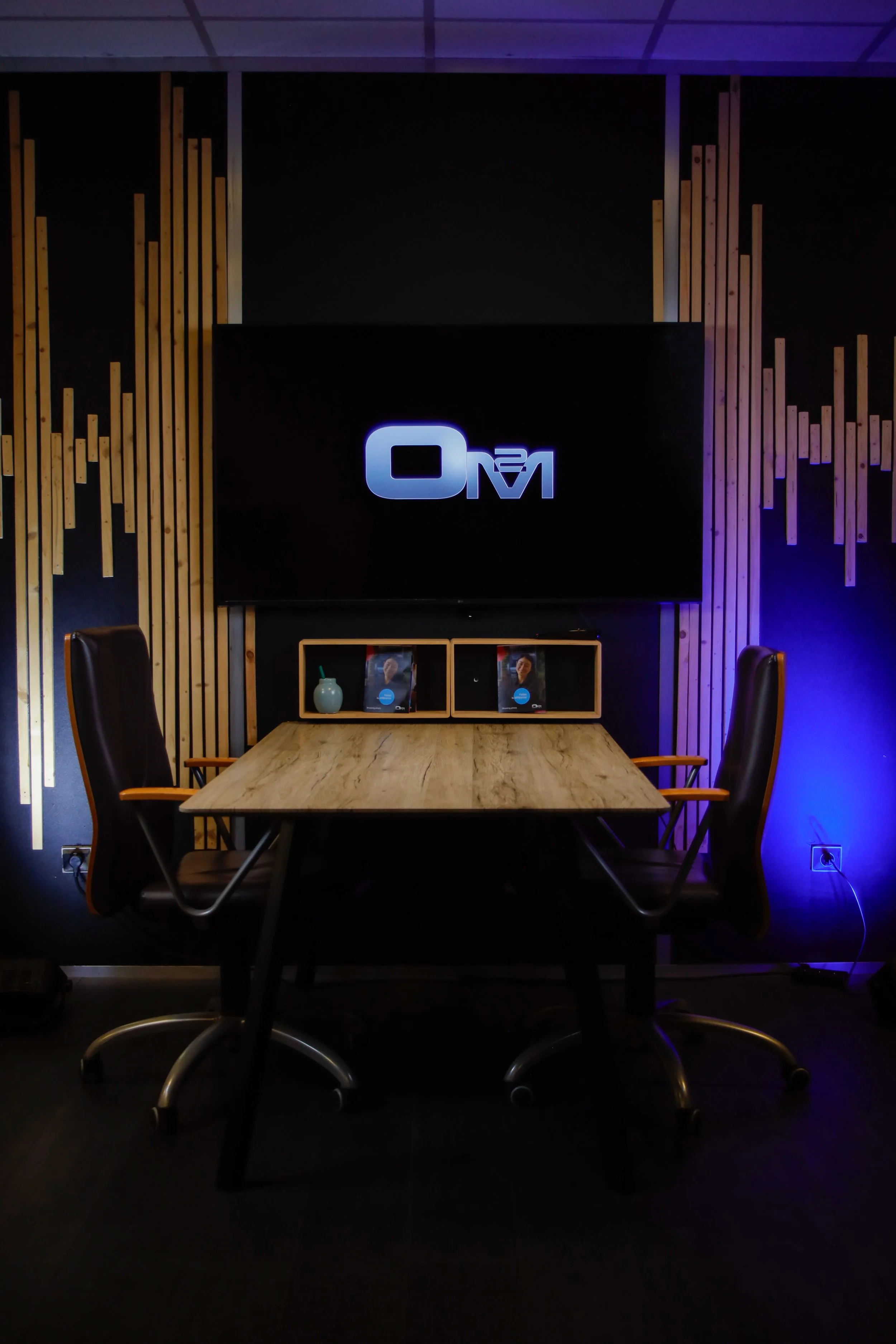 Salle de réunion avec un écran noir affichant un logo luminescent, deux chaises en cuir autour d'une table en bois, des décorations en bois sur le mur et un éclairage bleu à droite.