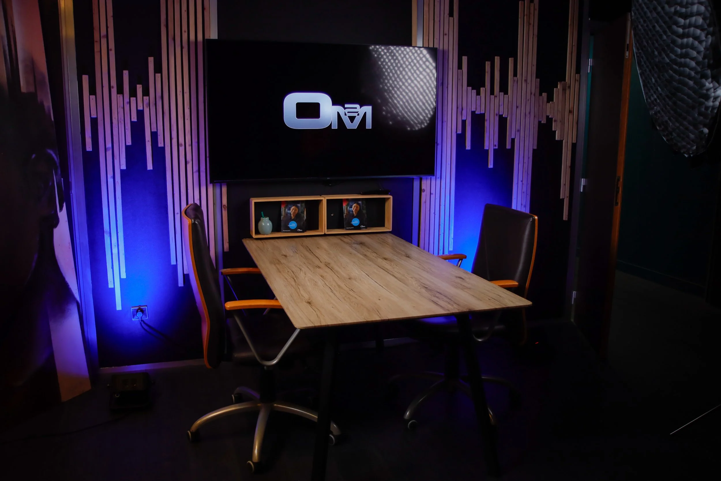 Salle de réunion avec table en bois, deux chaises de bureau, écran avec logo 'O M' et ambiance lumineuse bleue.