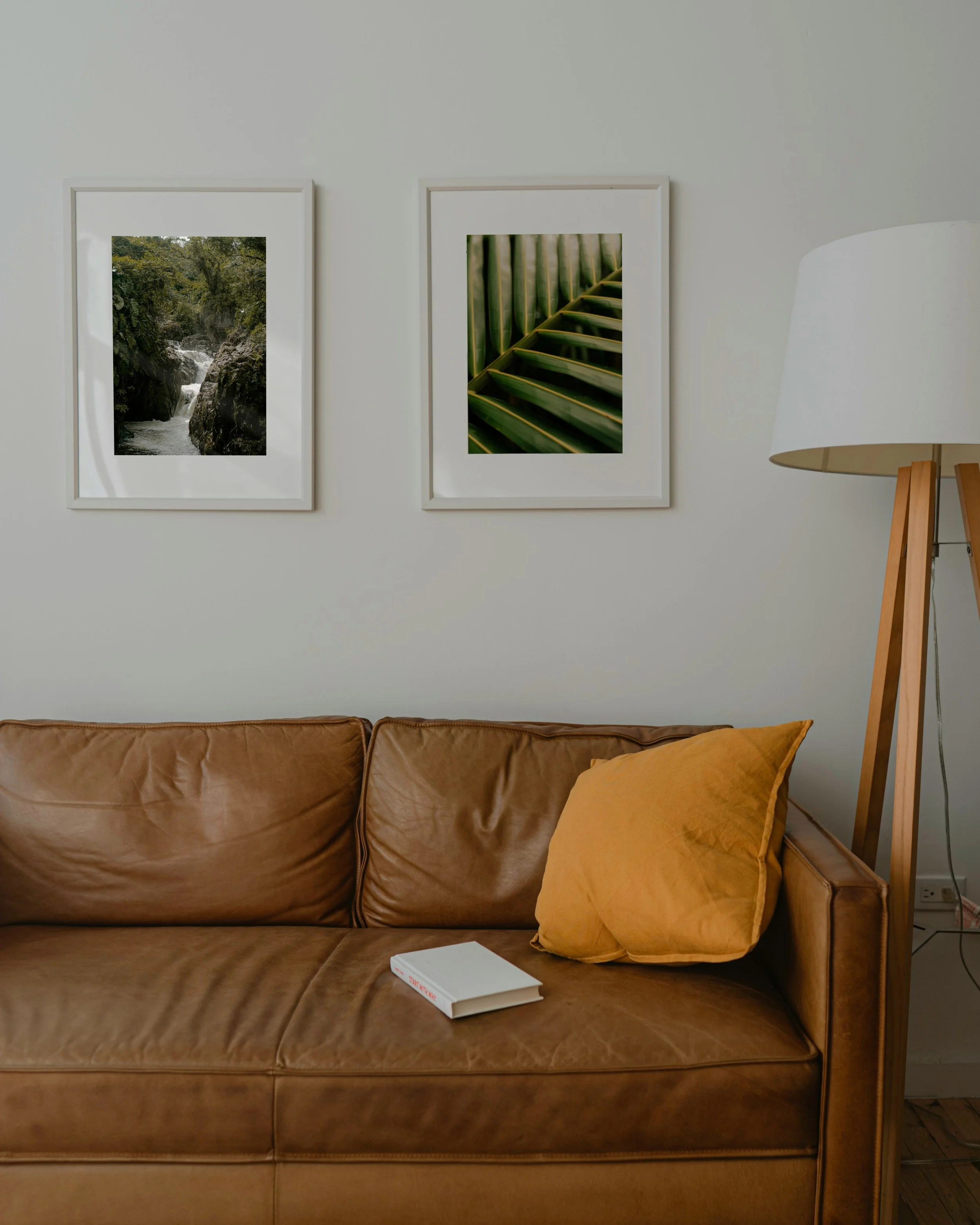 A brown leather couch with a mustard-colored pillow on the right side. A white book is placed on the couch in the lower center. Behind the couch, there are two framed pictures on a white wall; the left picture shows a waterfall in a forest, and the right picture shows green foliage. To the right of the pictures, there is a floor lamp with a white shade and wooden tripod legs.