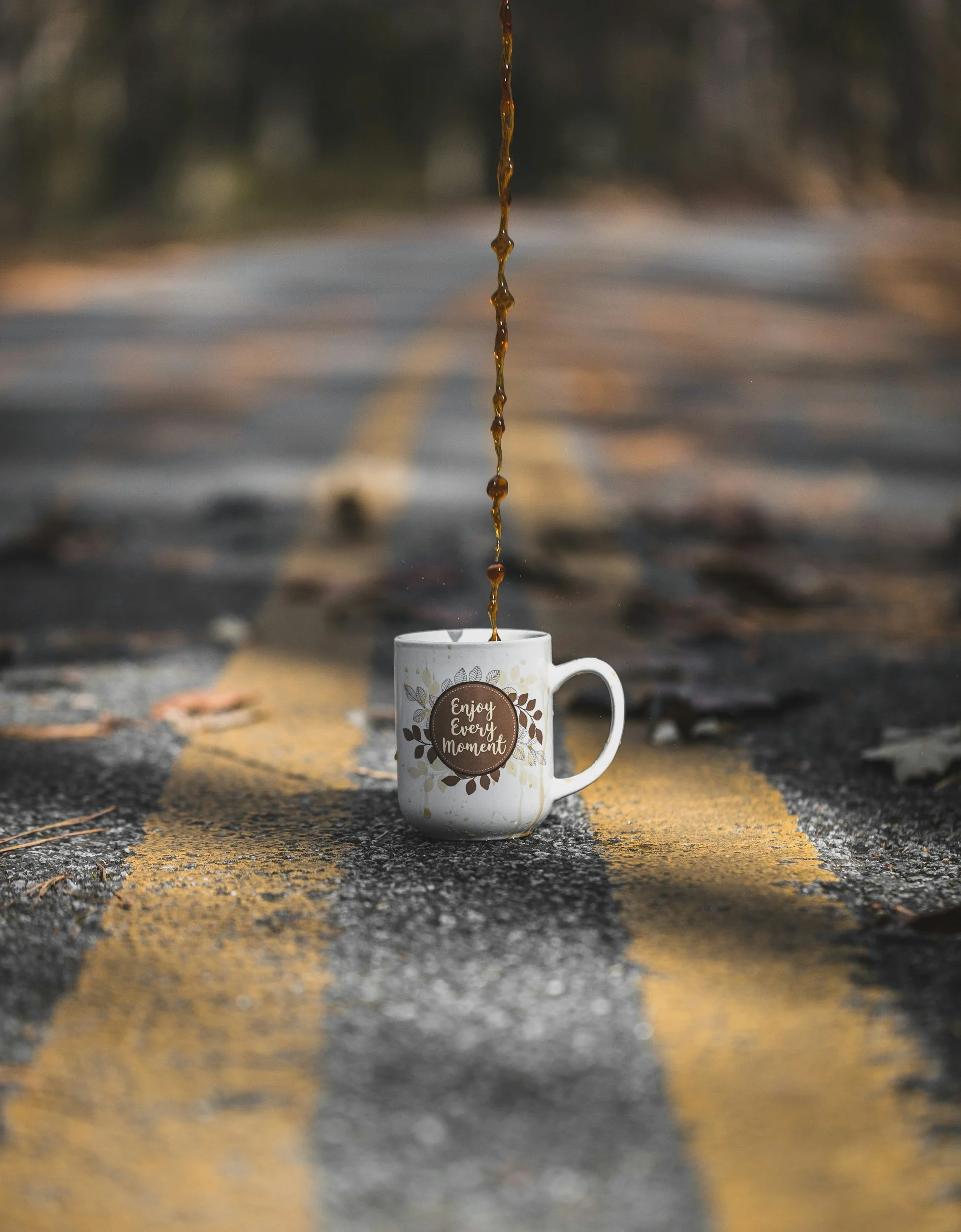 A white coffee mug with the quote 'Enjoy Every Moment' versus a road with a yellow line, a blurred background, and a stream of coffee pouring into the mug from above.