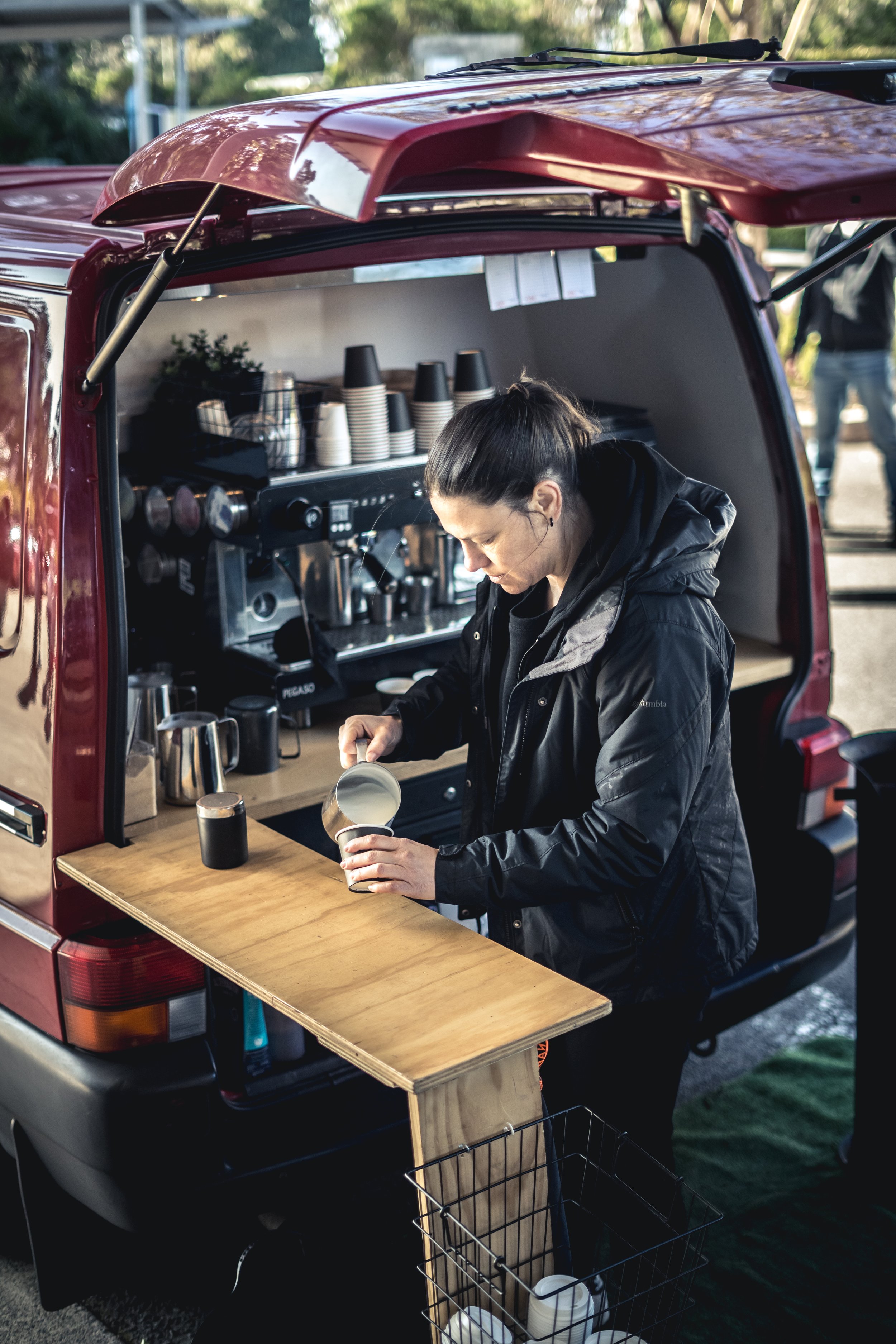 A woman making coffee from a portable coffee bar setup inside the back of a red SUV, with cups and coffee-making equipment visible.