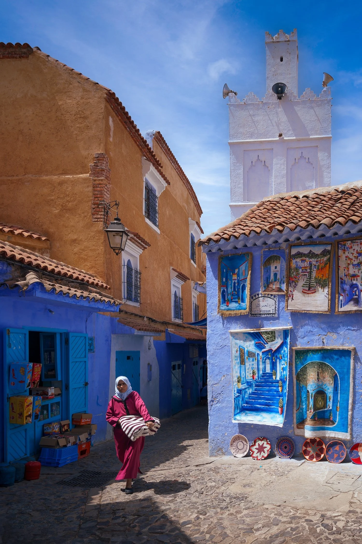 A woman in a pink outfit and white headscarf walking through a colorful street with blue shops and a display of painted ceramics, with buildings featuring orange and white walls and a tower in the background.