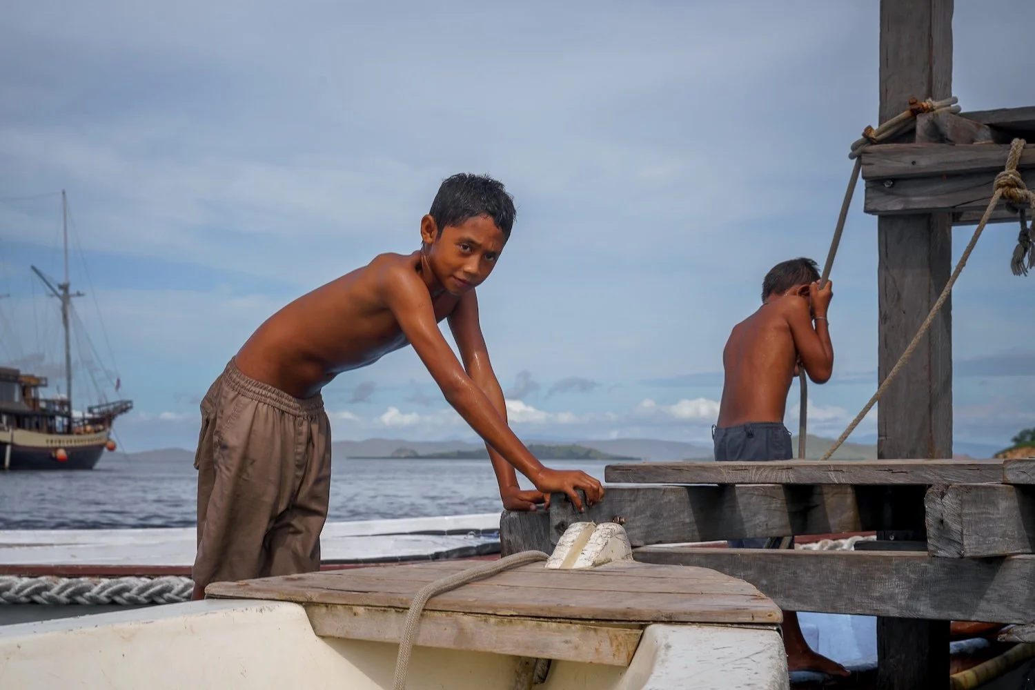 Two young boys with dark hair and dark skin, shirtless, are on a wooden dock near a body of water. One boy is leaning over, looking at the camera, and the other boy is turning away, holding onto a rope. There are boats in the background under a partl
