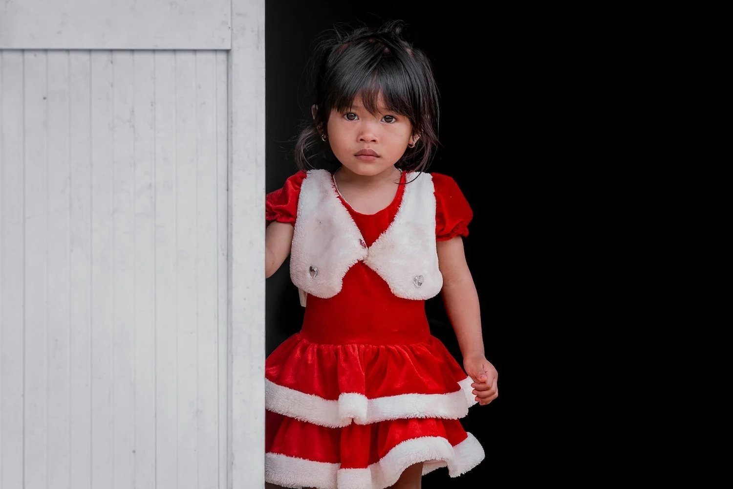 Young girl with dark hair wearing a red and white Christmas dress, standing partly behind a white wooden door, looking directly at the camera.