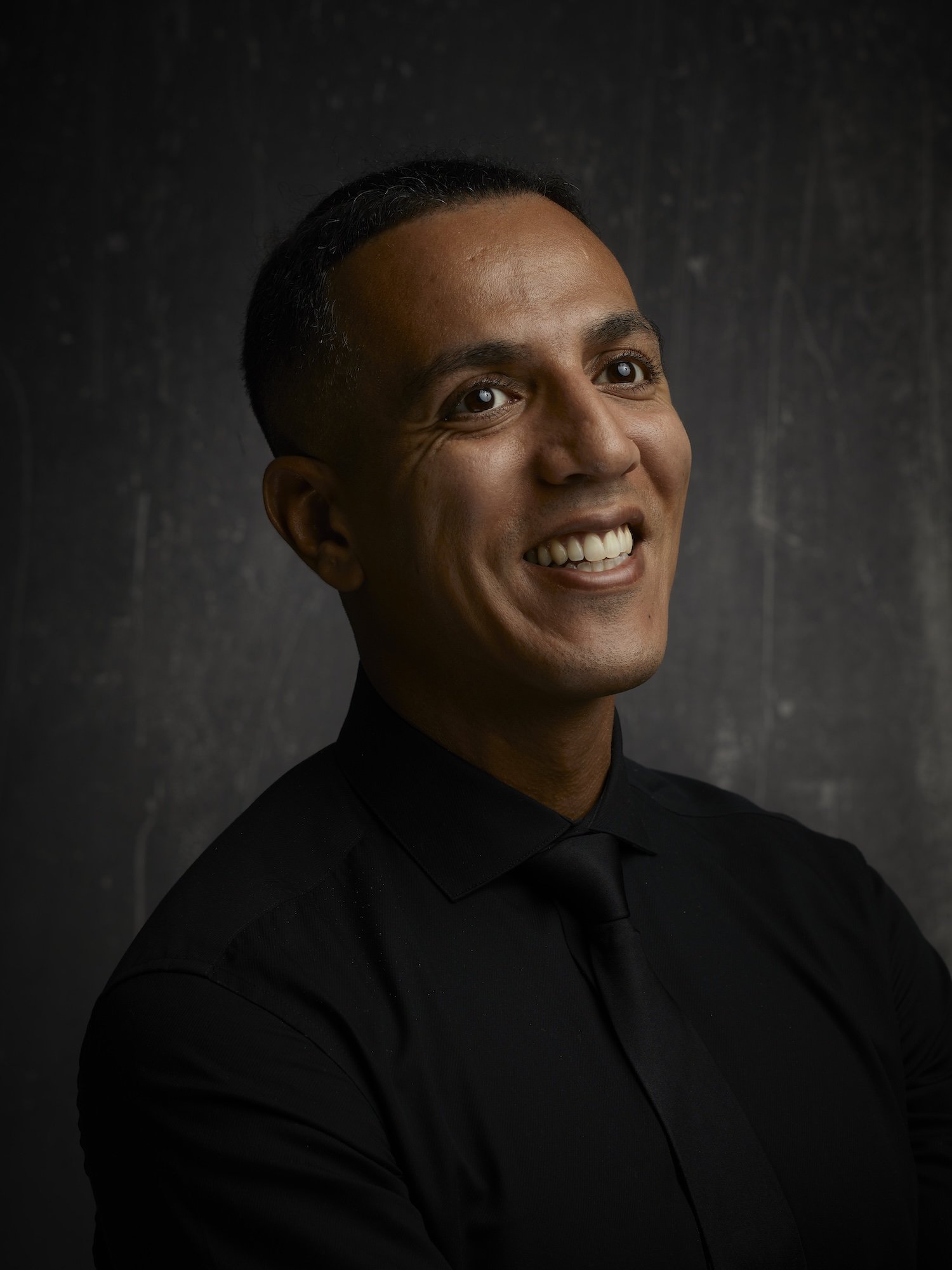 A portrait of a man smiling, wearing a black shirt, against a dark background.