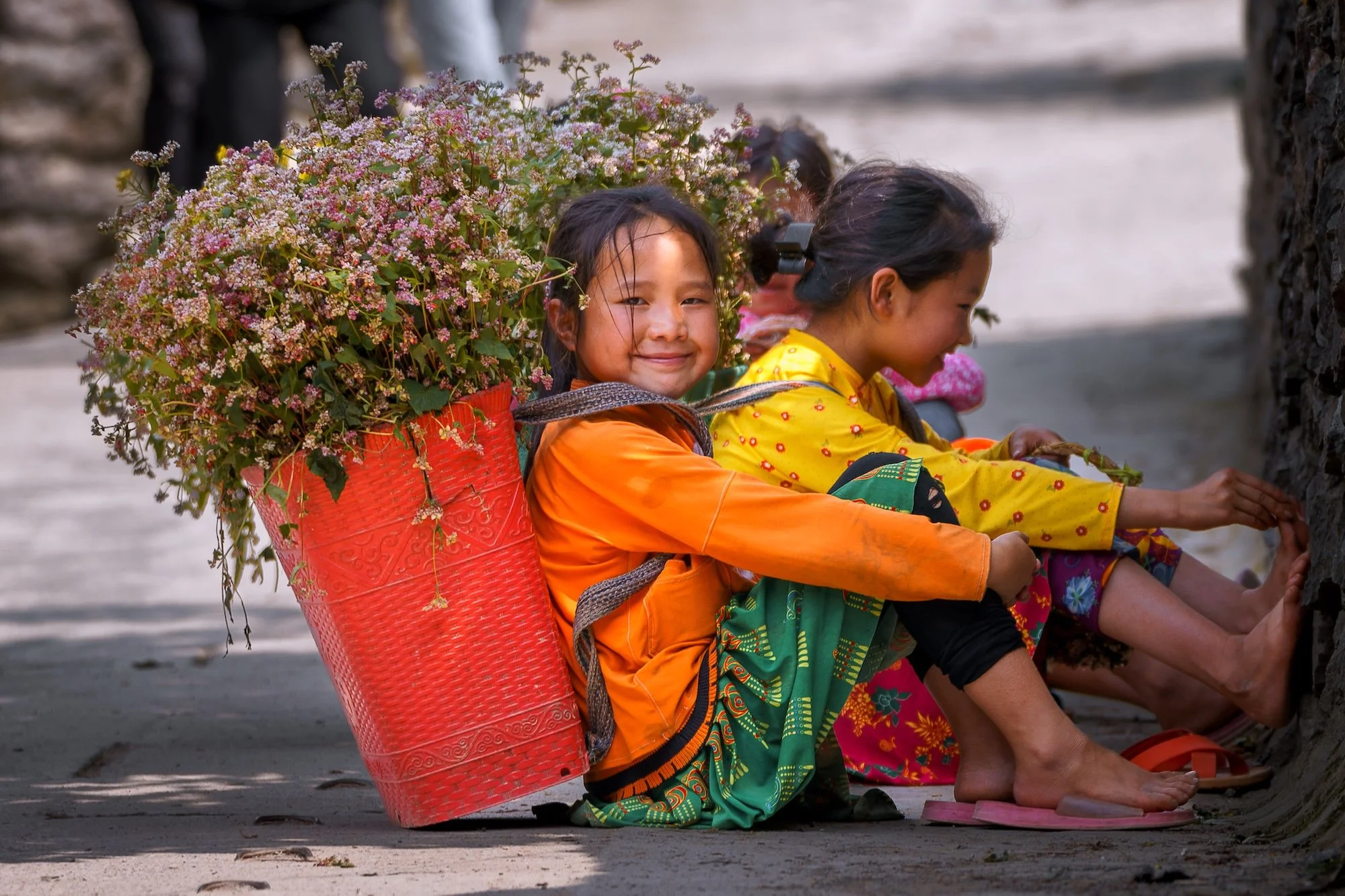 Group of children sitting on the ground, praying, with one girl smiling and carrying a large basket of pink and white flowers on her back.