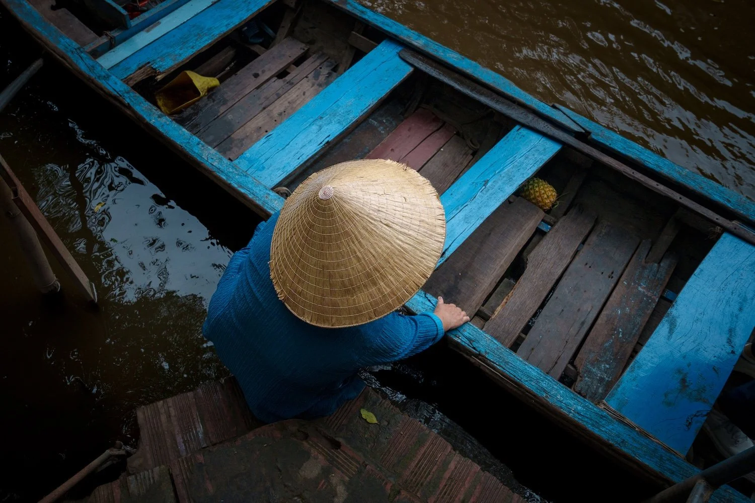 A person wearing a conical straw hat and a blue shirt is preparing to get into a wooden boat painted blue, with a pineapple visible inside. The boat is on a river or canal, and the person is stepping onto a wooden dock.