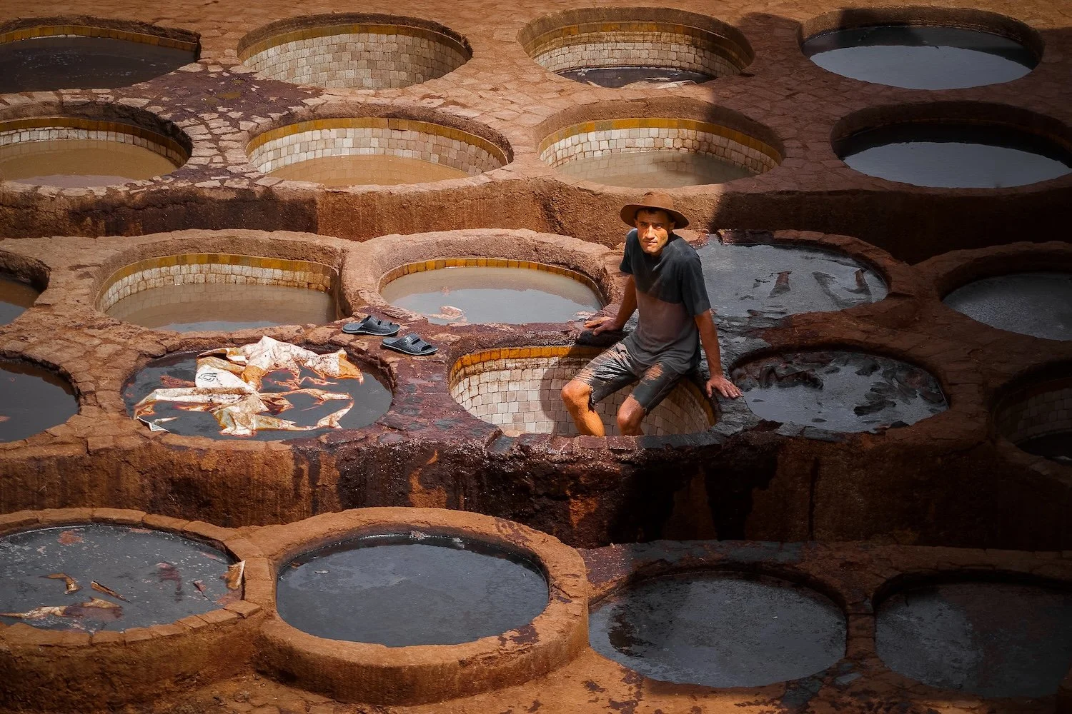 A man sitting on the edge of a large clay or brick construction with multiple circular open reservoirs filled with water. He is wearing a wide-brimmed hat, a dark shirt, and shorts, with his feet in the water. There are shoes and some cloth or paper 