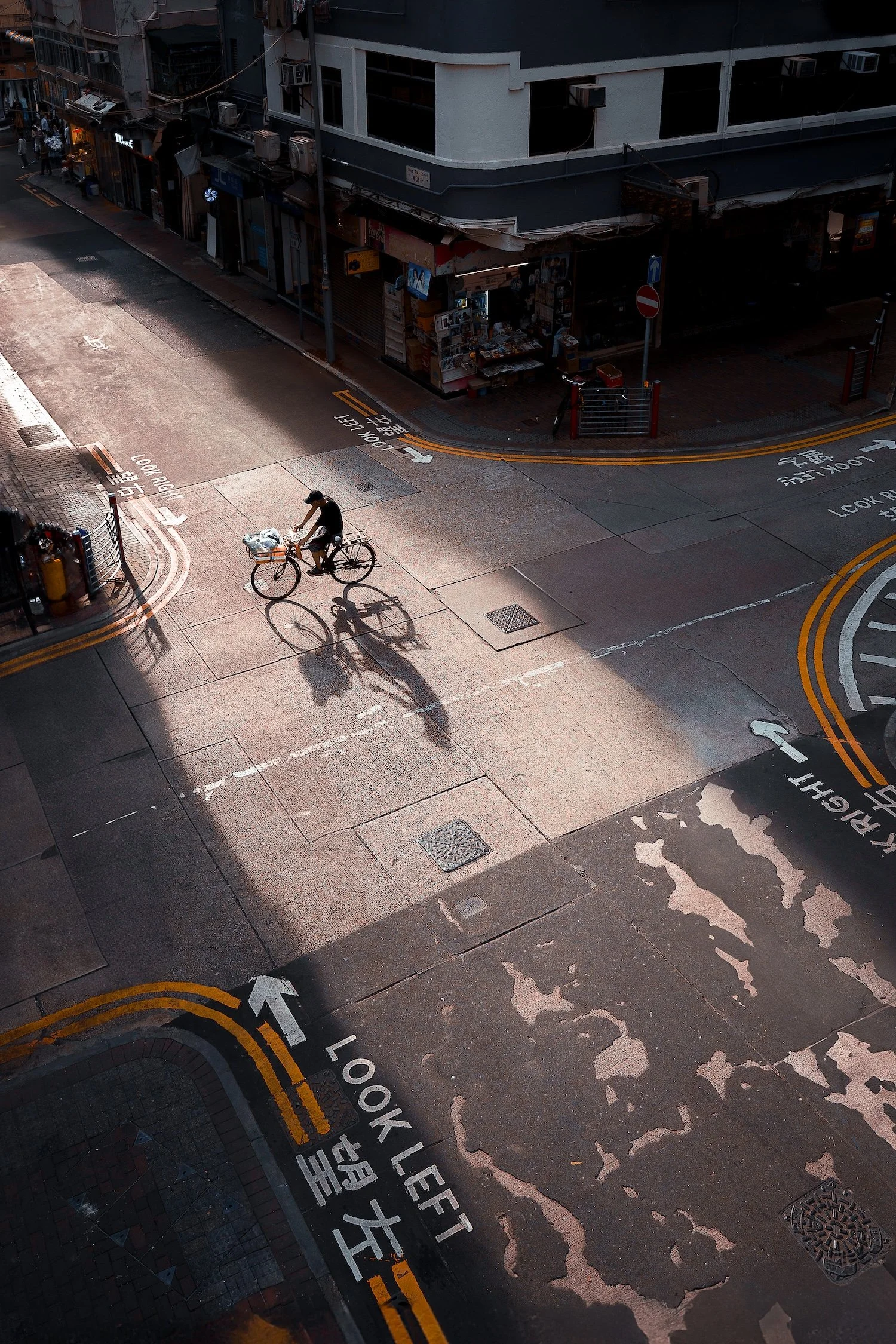 A person riding a bicycle on a city street during daytime, casting a long shadow, with buildings and storefronts in the background.