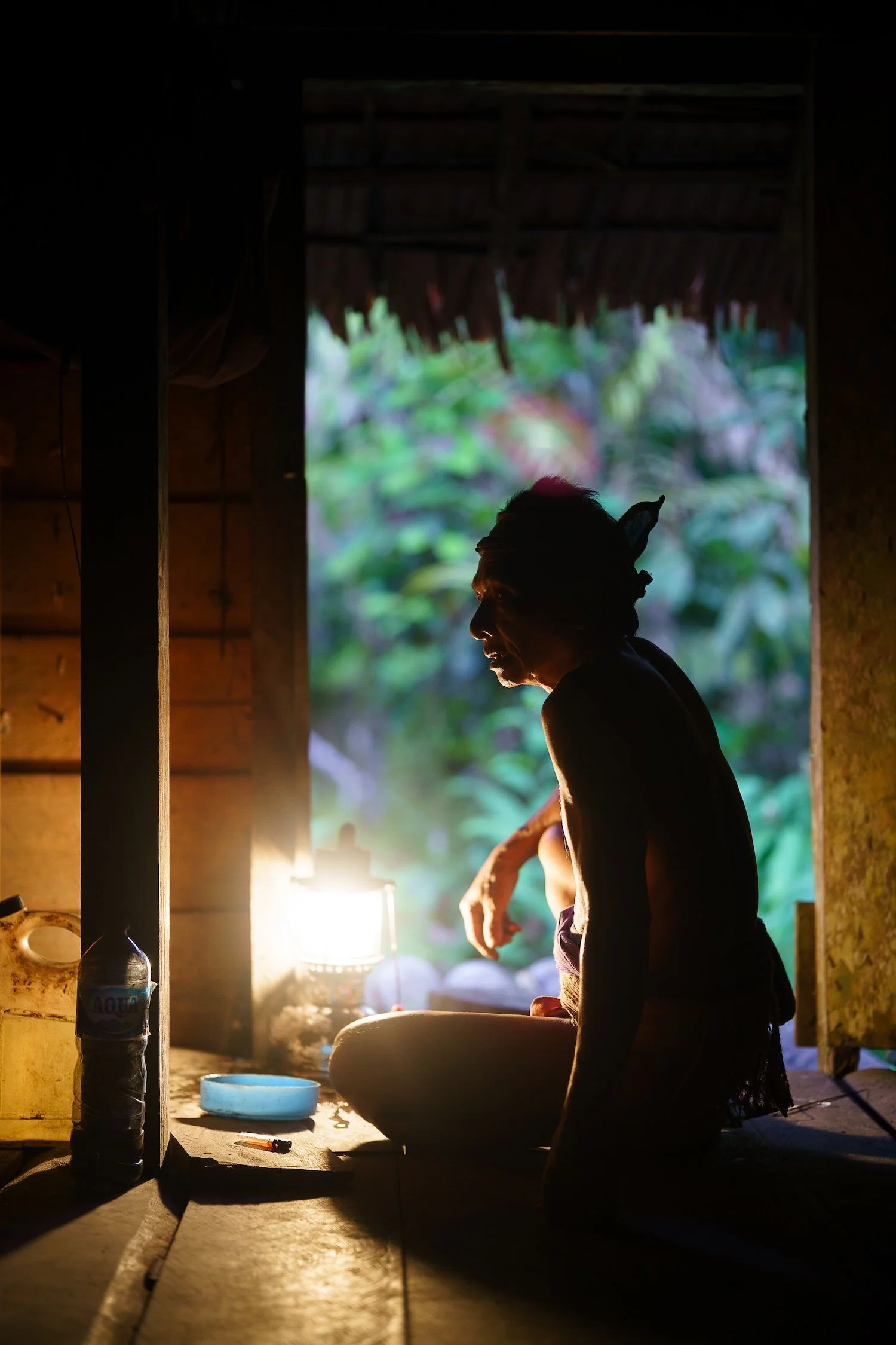 A person with long hair and a headband is sitting in a wooden hut, with lush green foliage visible outside. The scene is backlit, creating a silhouette of the person. Items like a water bottle, a bowl, and a lit lantern are on the table inside.