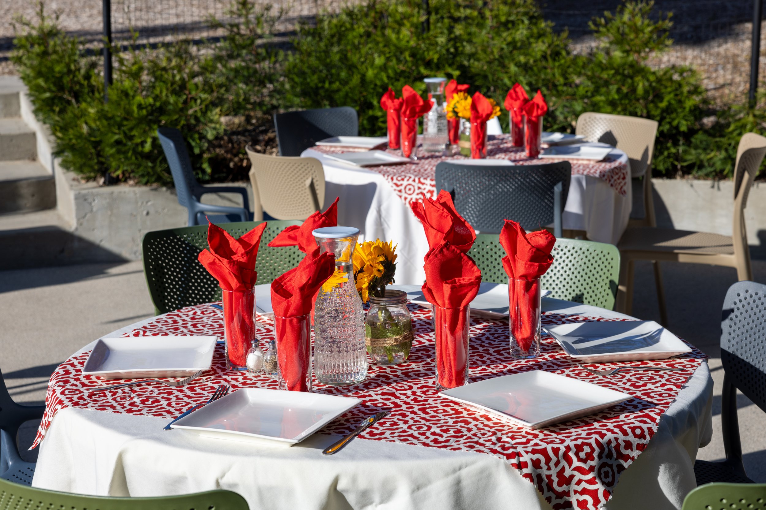 Outdoor dining tables set for a wedding reception meal, decorated with red napkins in glasses, bottles of water, flowers, and white plates, surrounded by chairs and greenery.