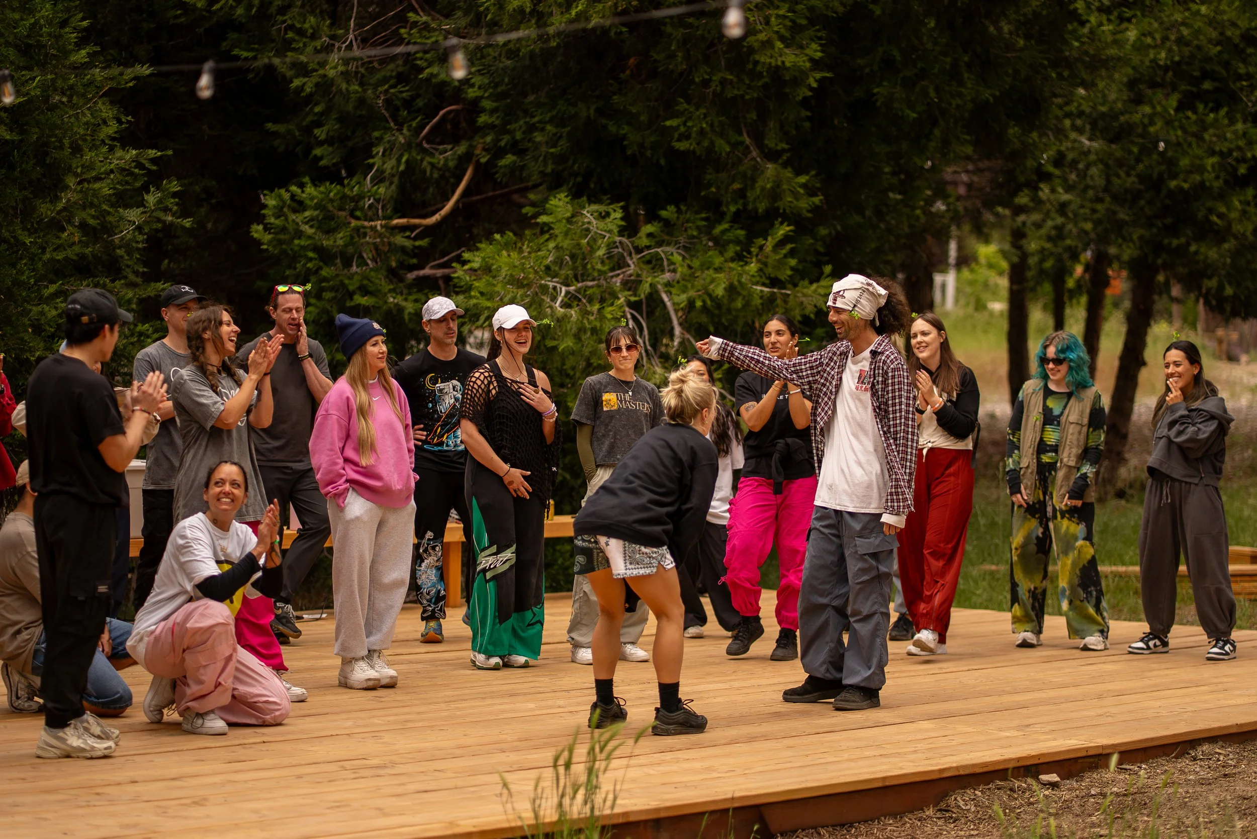 People practicing yoga or meditation on a wooden deck outdoors in a forested area, with trees and greenery surrounding them.