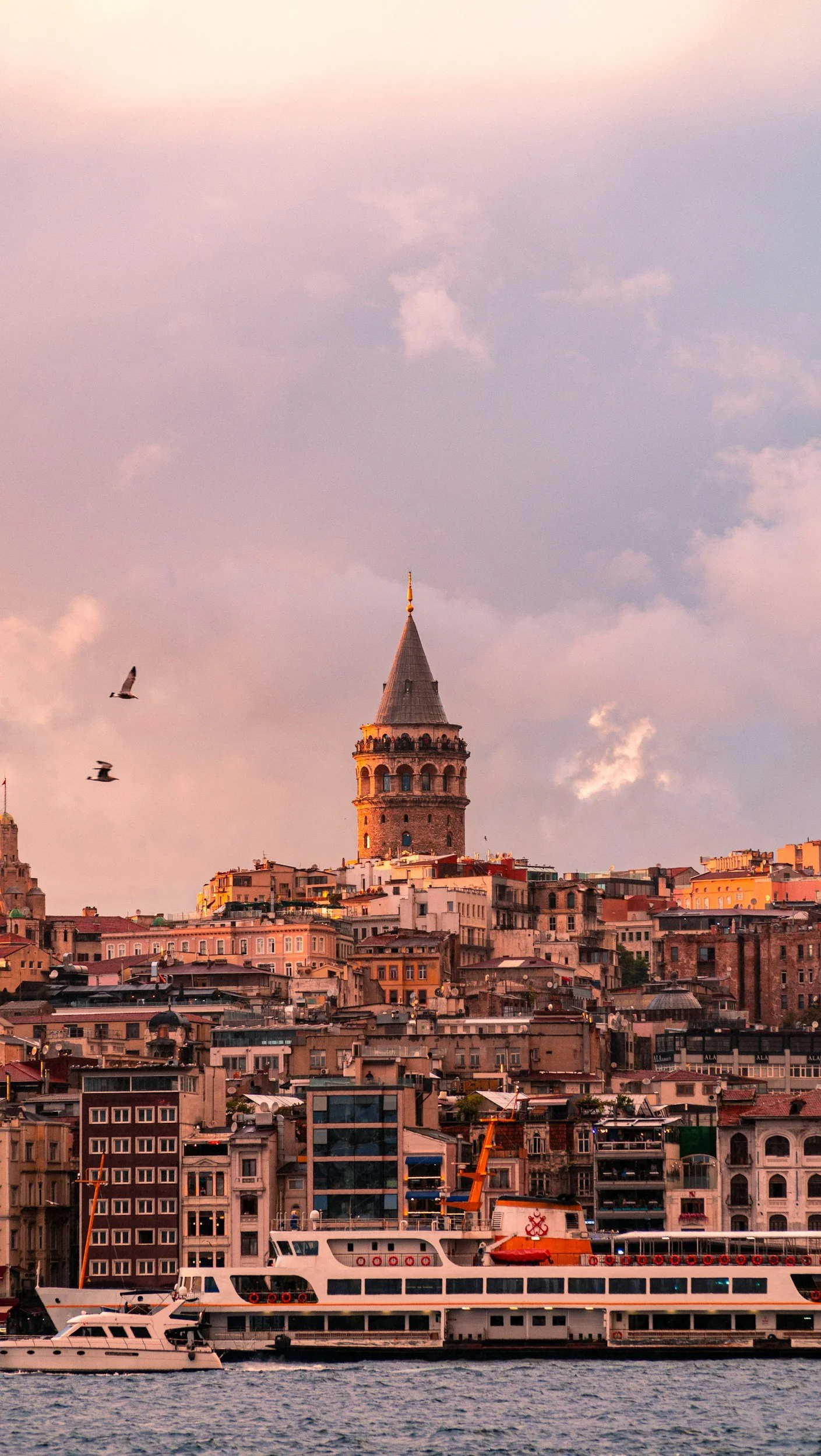 A city skyline with a historic tower, boats in the Water, and a pink and purple sky at sunset.