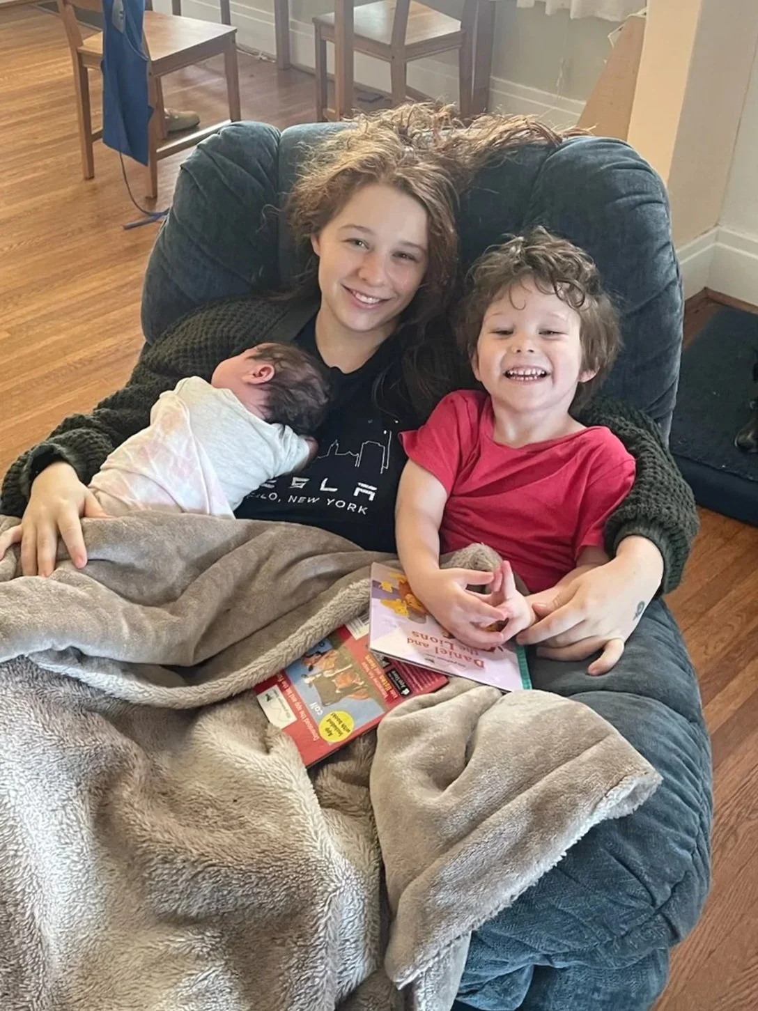 A woman with curly hair and two children sitting on a couch, one child is a newborn resting on her chest and the other is an older boy smiling at the camera, all smiling and happy.