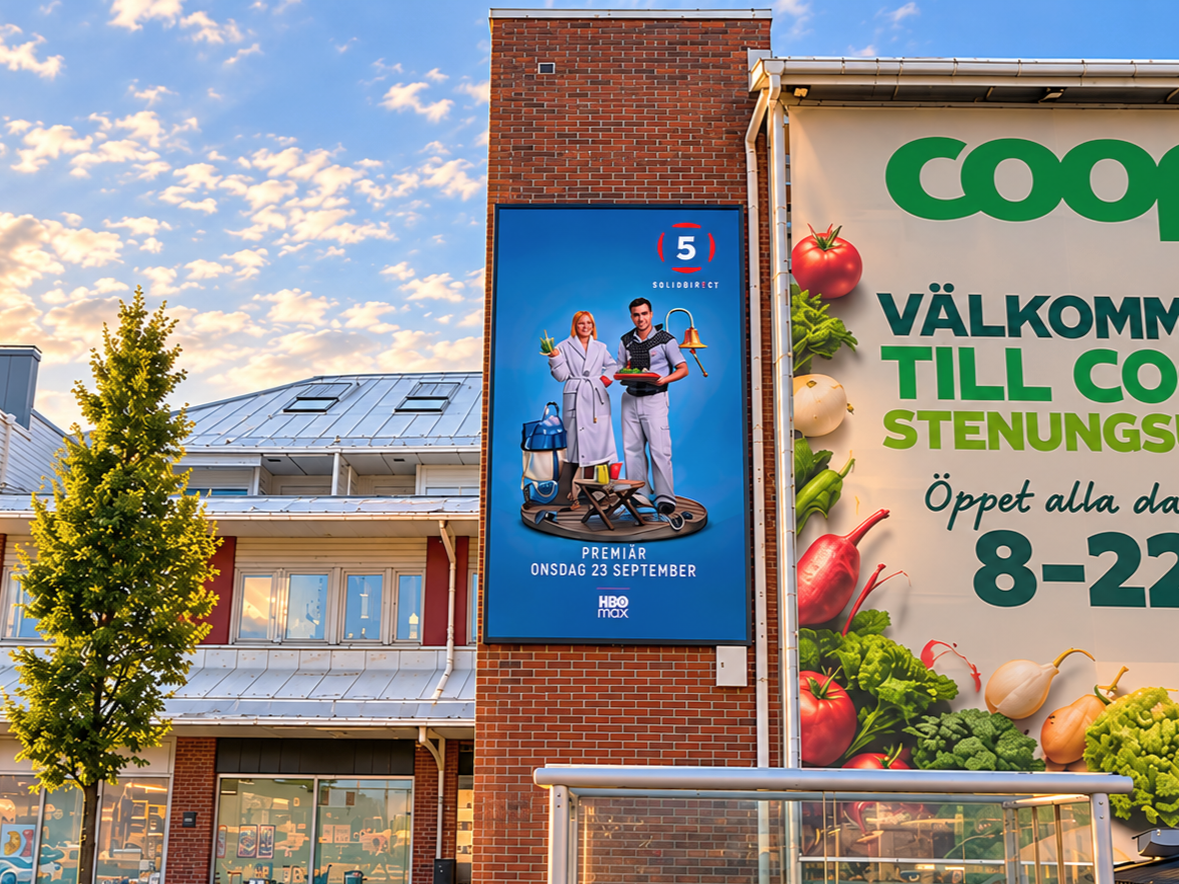 Coca-Cola reklamskylt till vänster, toalettstängsel och ingång till COOP stormarknad till höger, samt en bilparkering framför.