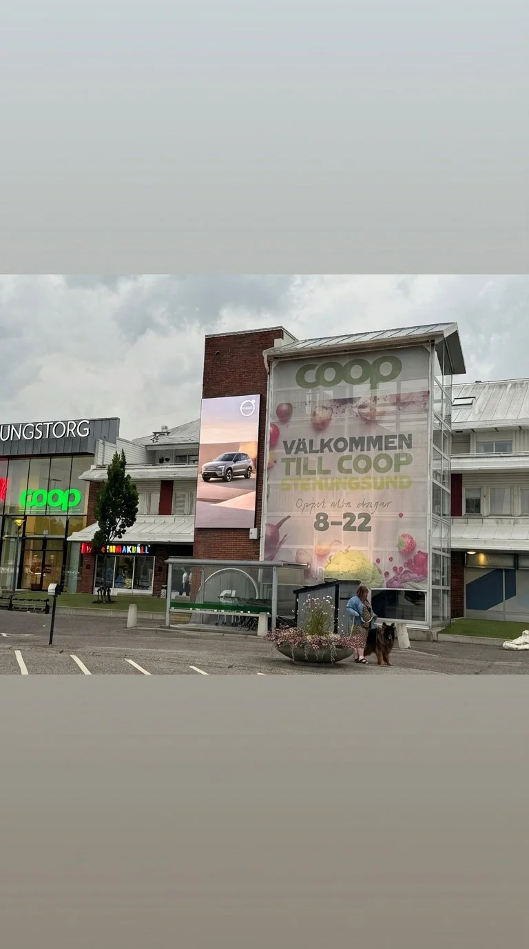 Coca-Cola reklamskylt till vänster, toalettstängsel och ingång till COOP stormarknad till höger, samt en bilparkering framför.