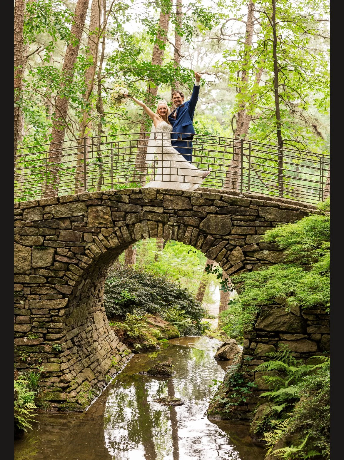 A bride and groom celebrating on a stone bridge over a creek in a lush green forest, holding hands and raising their arms in victory.