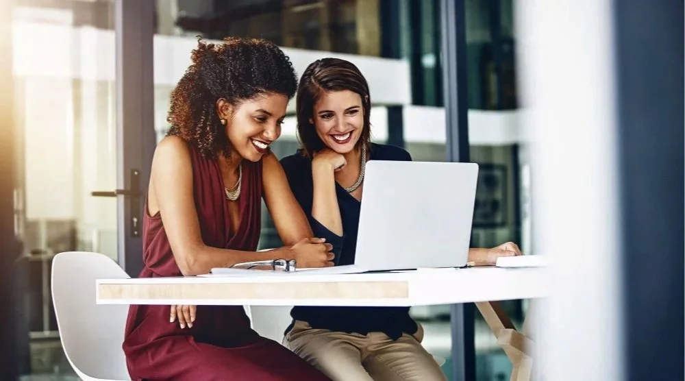 Two women smiling and looking at a laptop screen in an office setting.