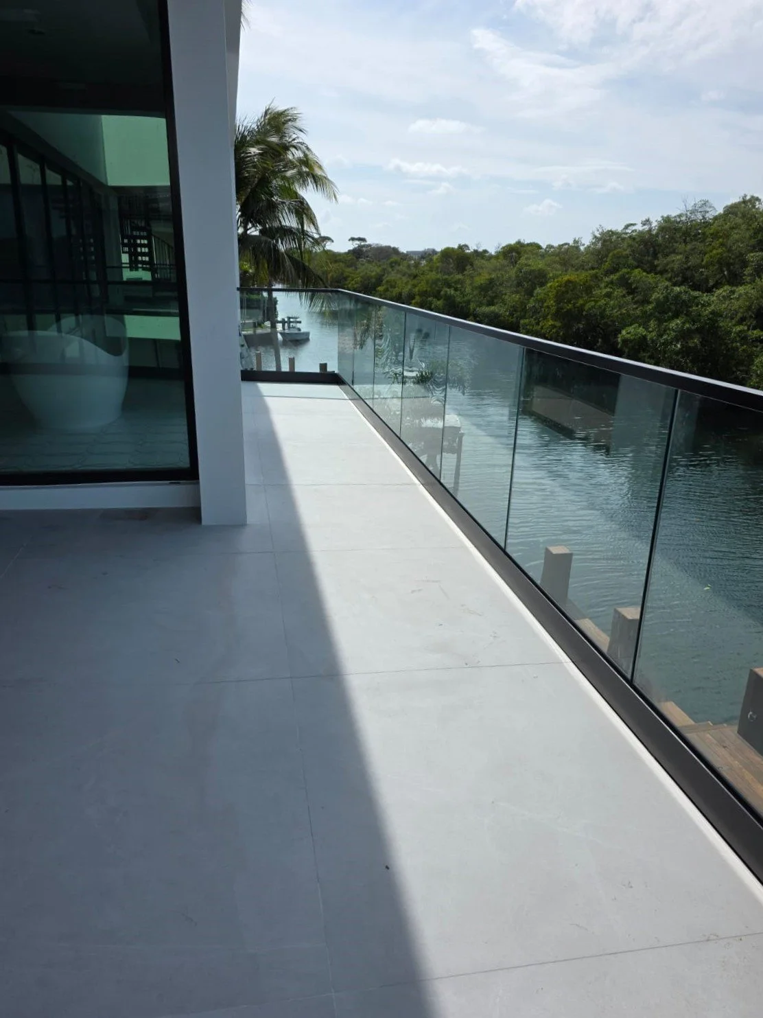 Modern balcony with glass railing overlooking a waterway and lush green trees, part of a contemporary building with large glass doors and white tiled flooring.
