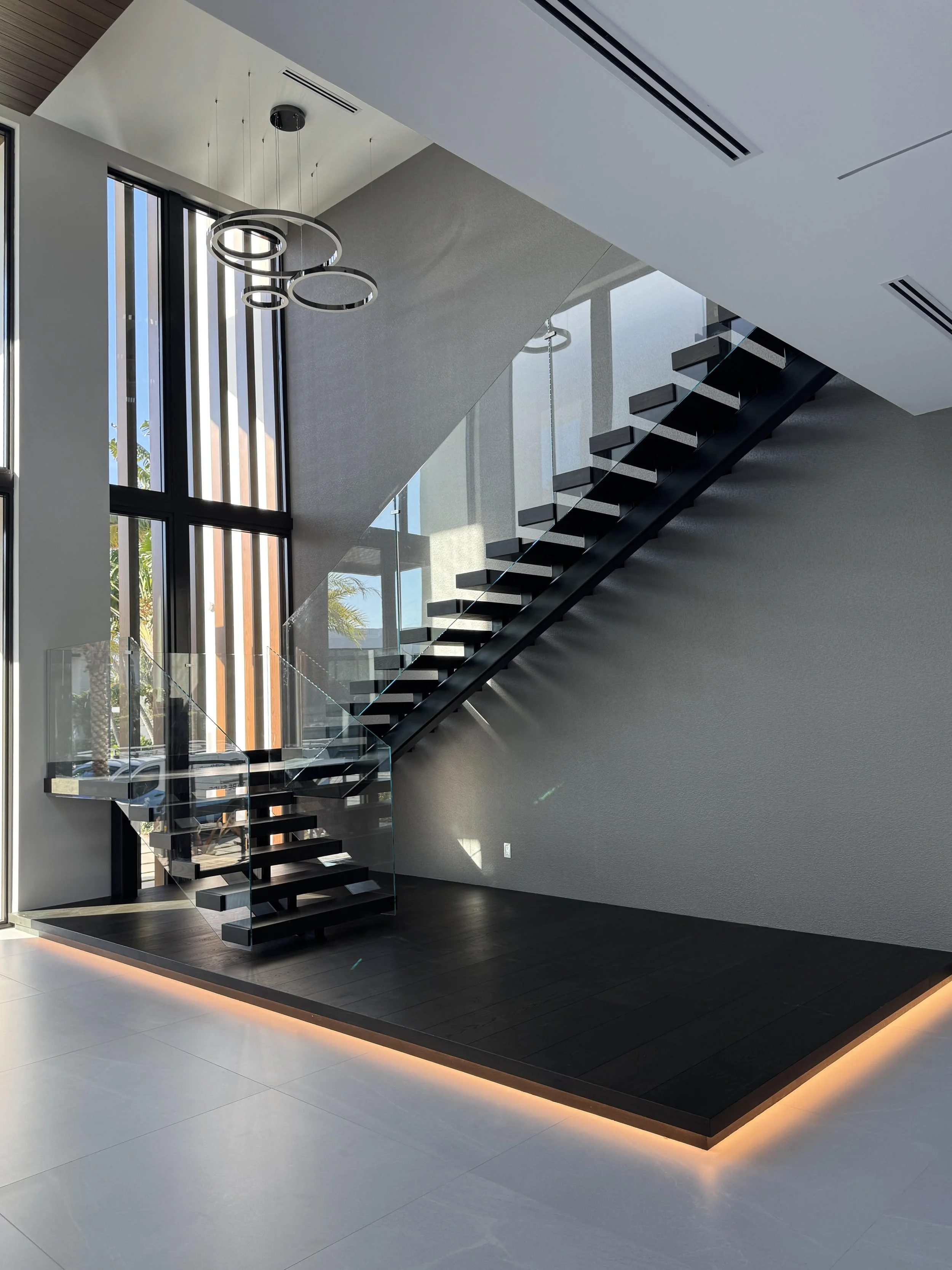 Modern staircase with open black steps, glass railing, located in a spacious area with large windows and high ceiling featuring a circular hanging light fixture, illuminated by natural daylight.