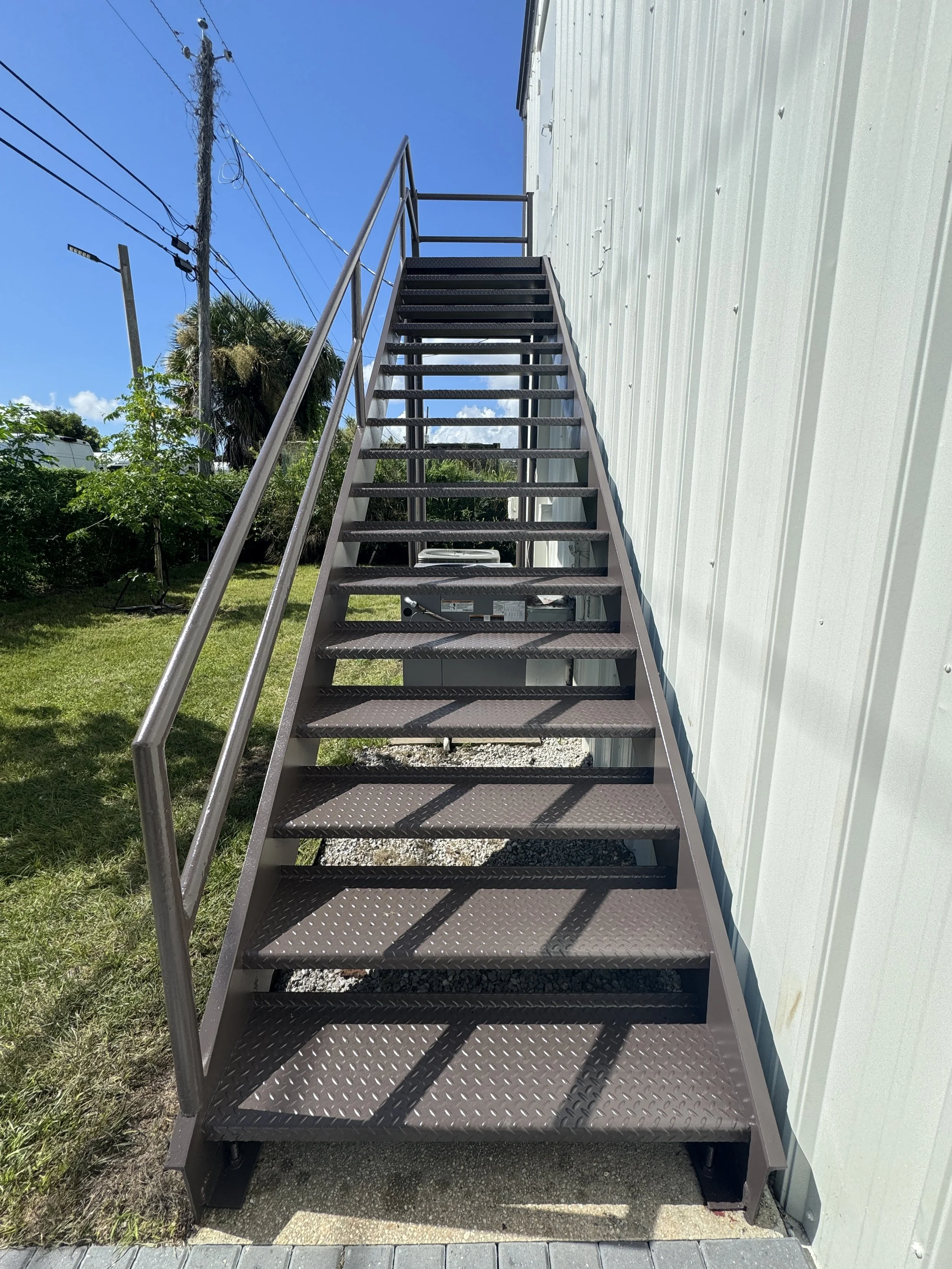 Metal outdoor staircase with handrails ascending beside a white metal building under a clear blue sky, with electric poles and foliage visible in the background.