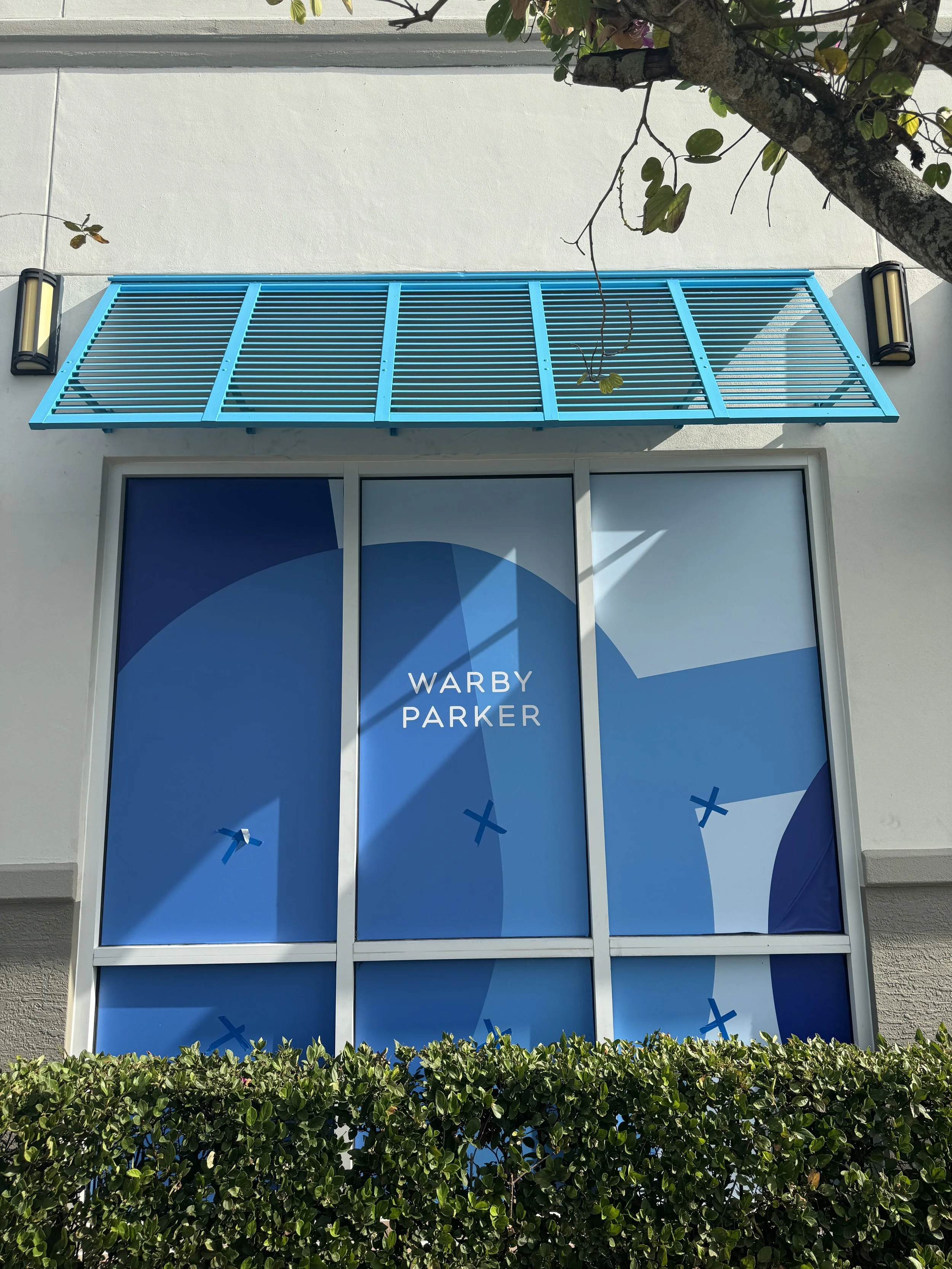 Blue window awning with horizontal slats, above a large window displaying the text 'WARBY PARKER' on a blue background, with a green hedge in front and two exterior wall-mounted light fixtures on either side of the window, all against a white wall wi