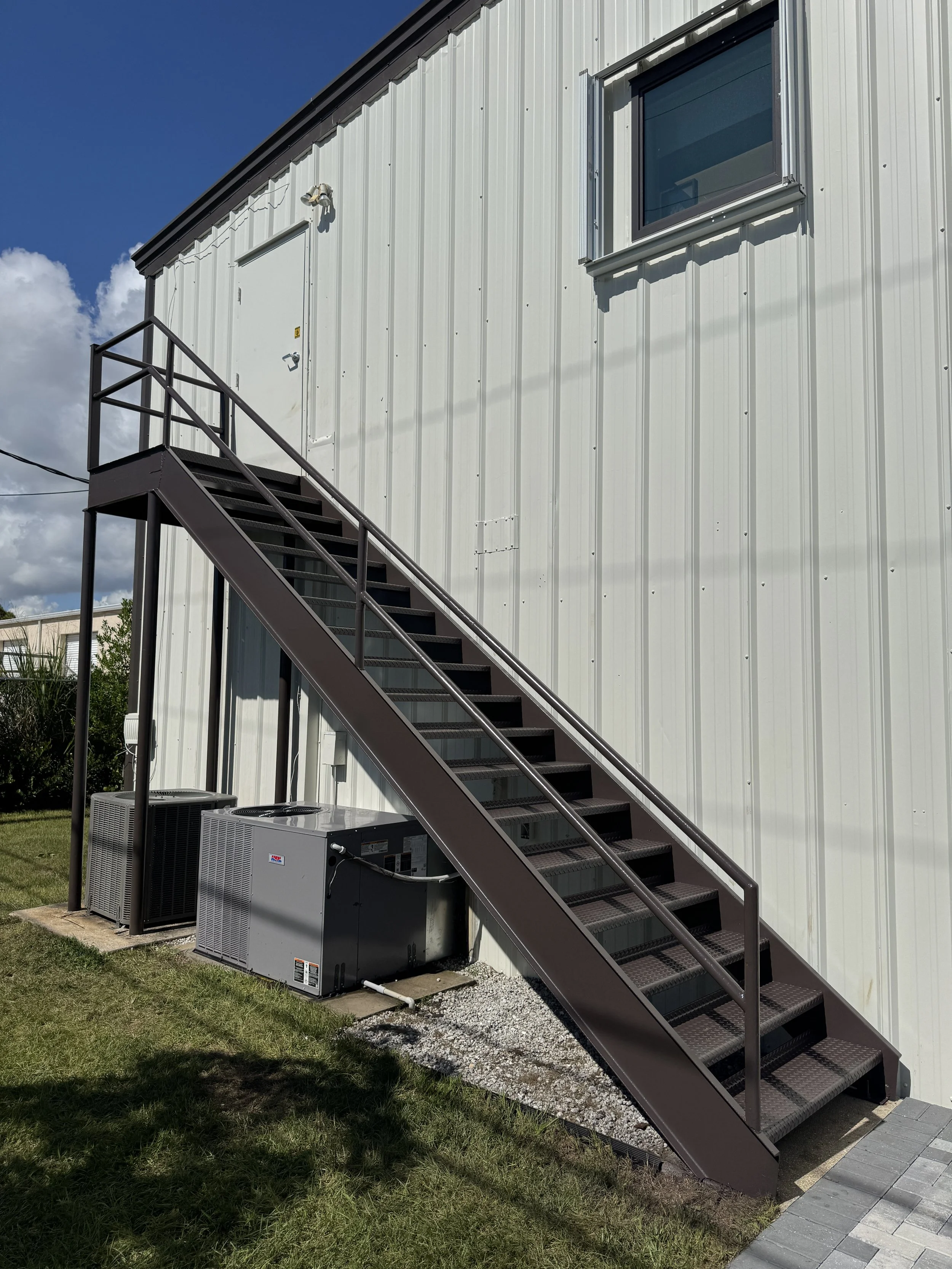 Exterior of a building with metal siding, a set of metal stairs with handrails leading to a door, and two HVAC units at the base of the stairs, under a partly cloudy sky.