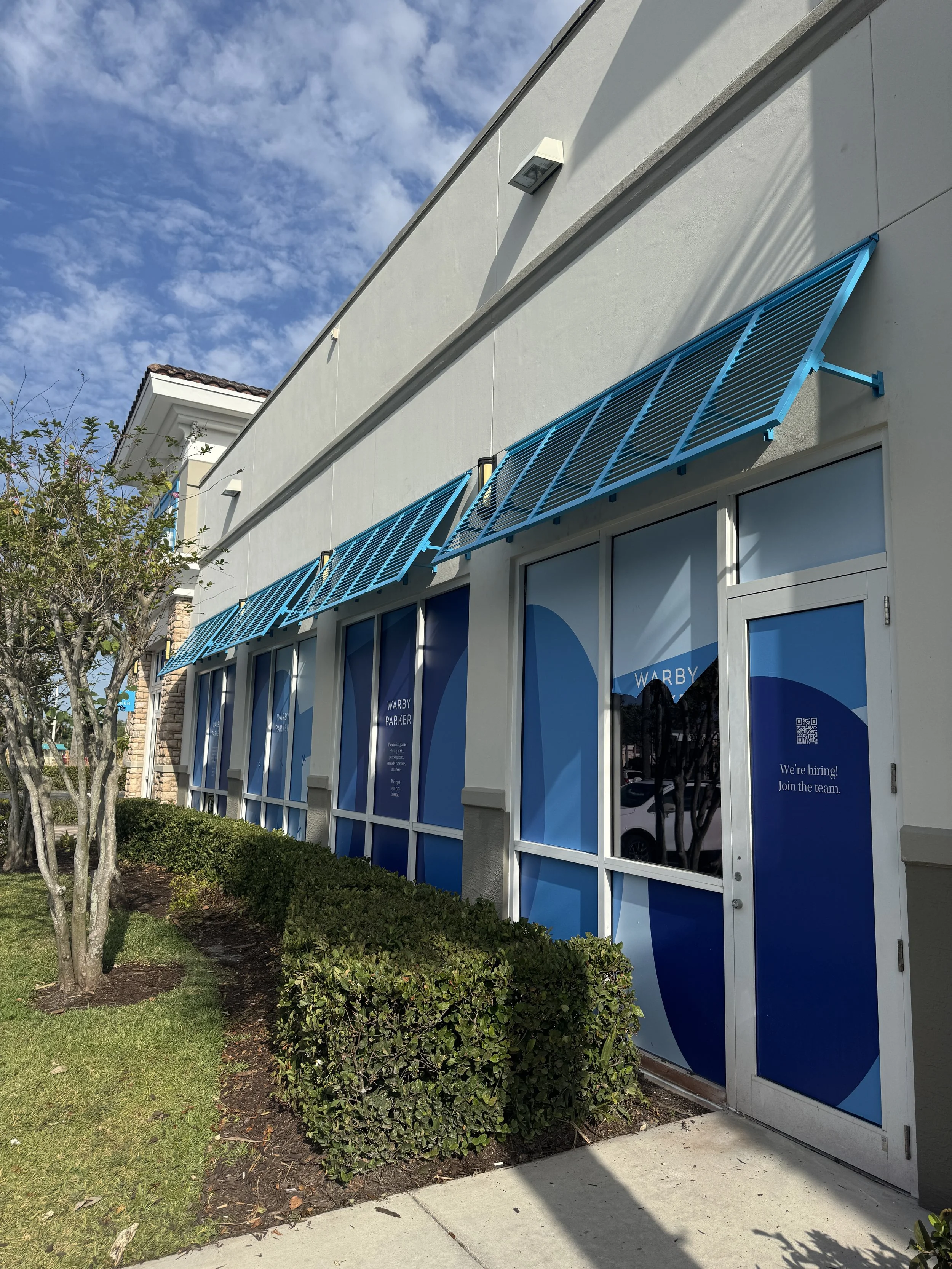 Storefront with blue awnings and posters that say 'Warby Parker' and 'We're hiring! Join the team.' A tree and bushes are in front of the store, and the sky is partly cloudy.