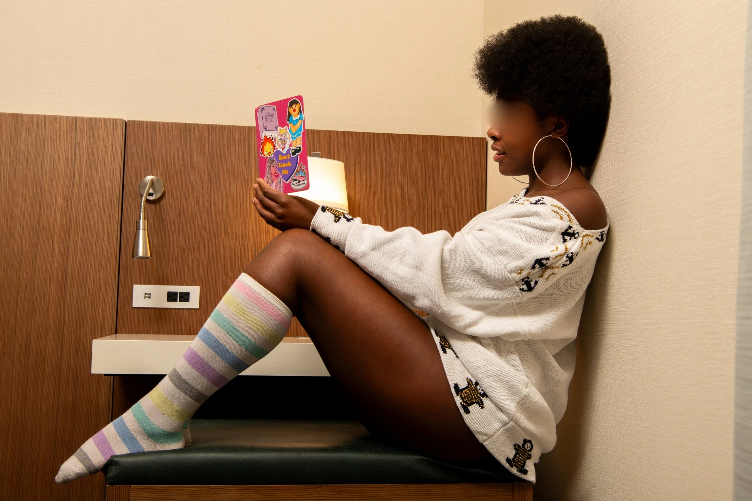 A woman with natural hair, wearing a white sweater with dog patterns, striped pastel knee-high socks, and large hoop earrings, is sitting on a hospital bed, reading a colorful card.
