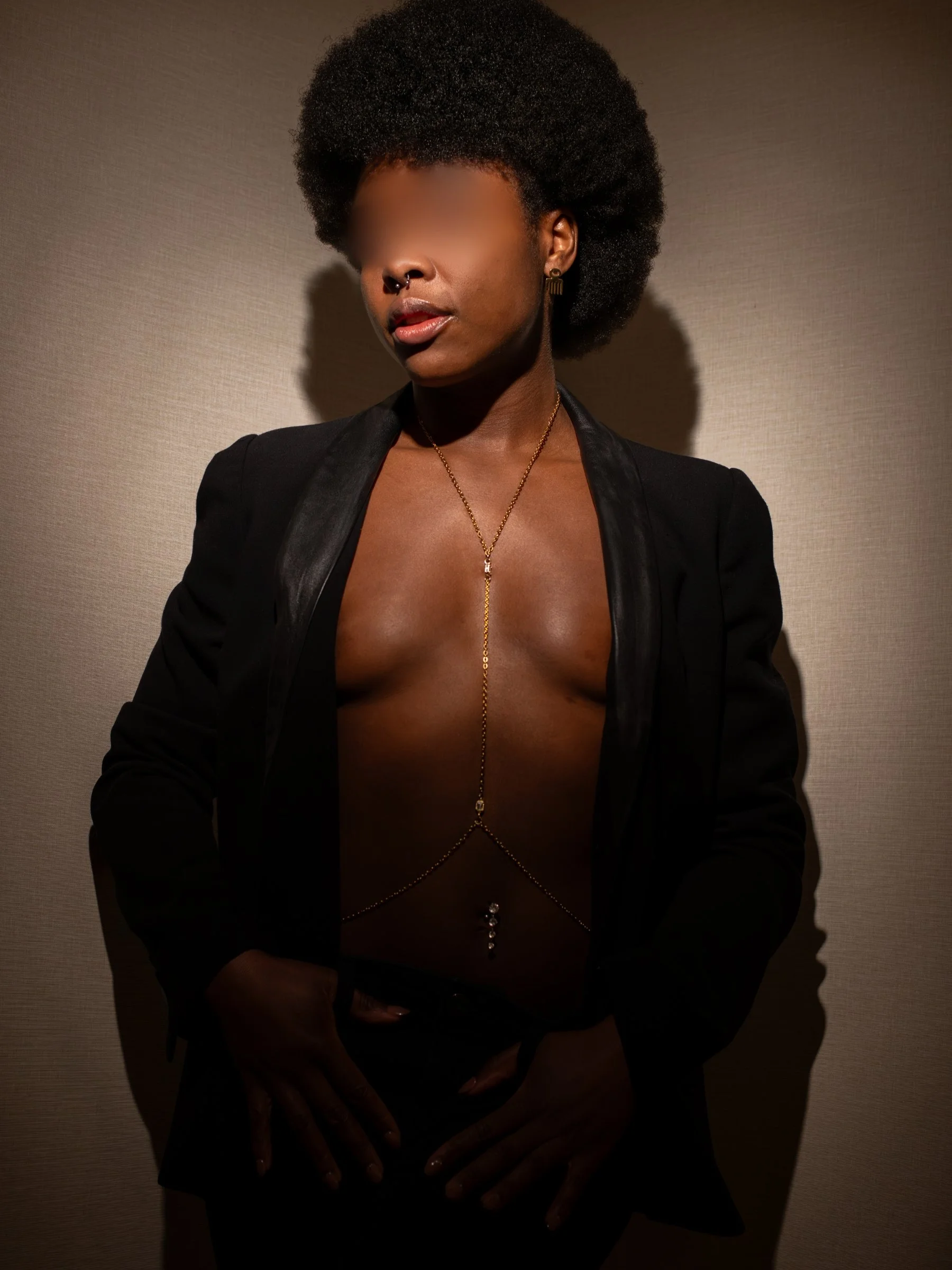 A female escort wearing gold body jewelry and a black jacket with moody lighting